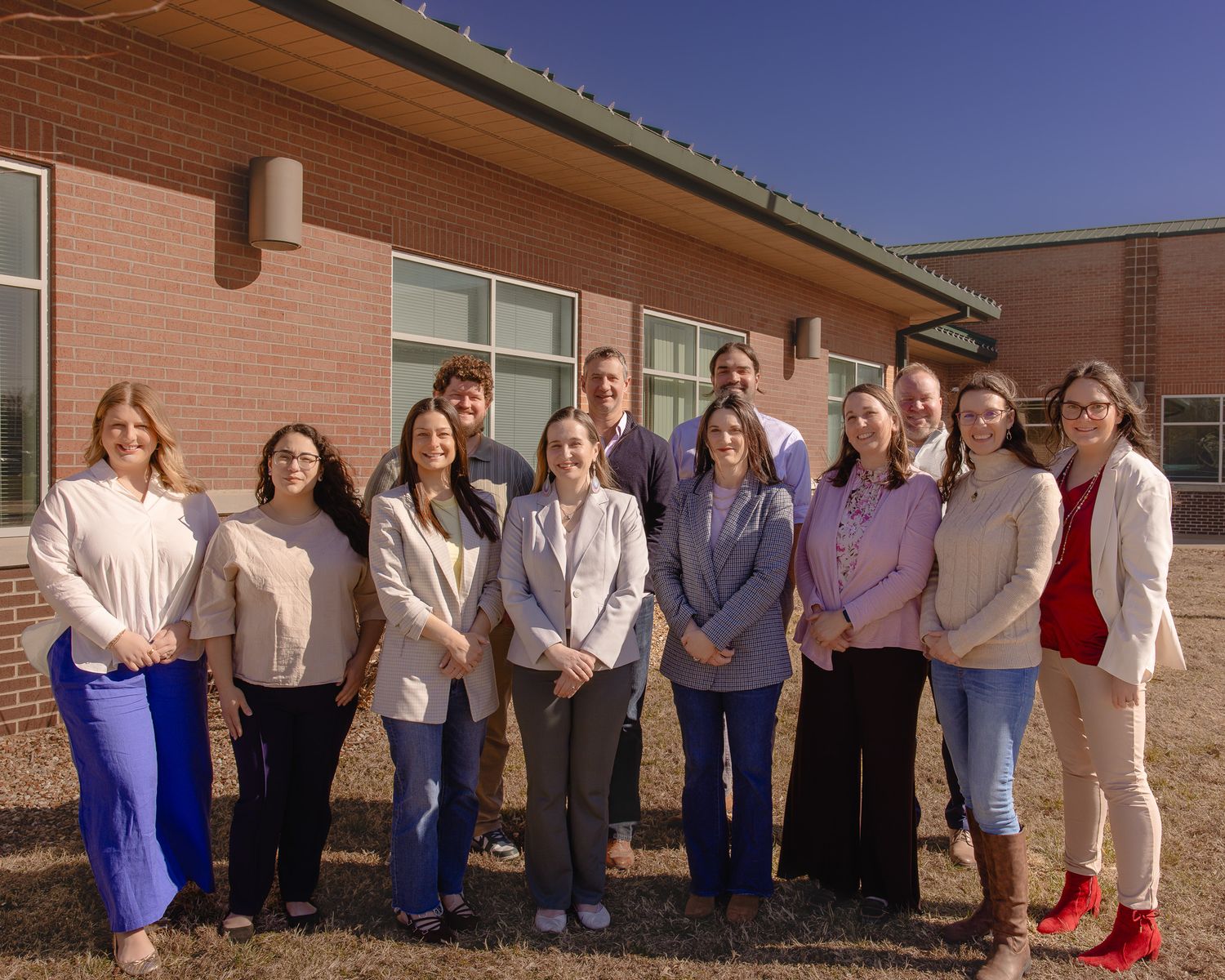 Group of healthcare professionals posing outdoors in front of a brick building on a sunny day.