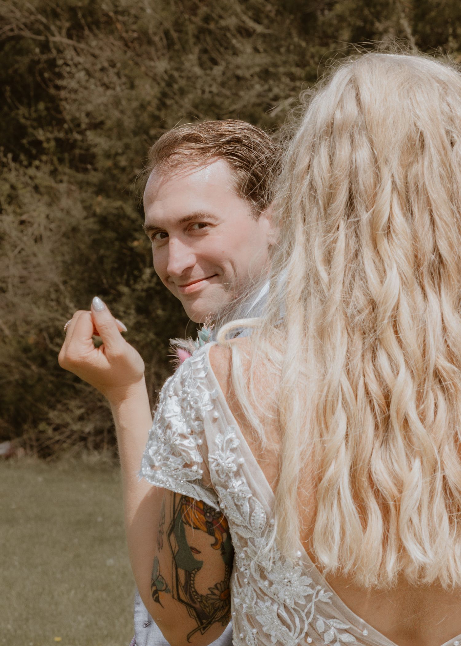 Groom with tattoos smiling at bride in lace wedding dress during outdoor wedding ceremony.