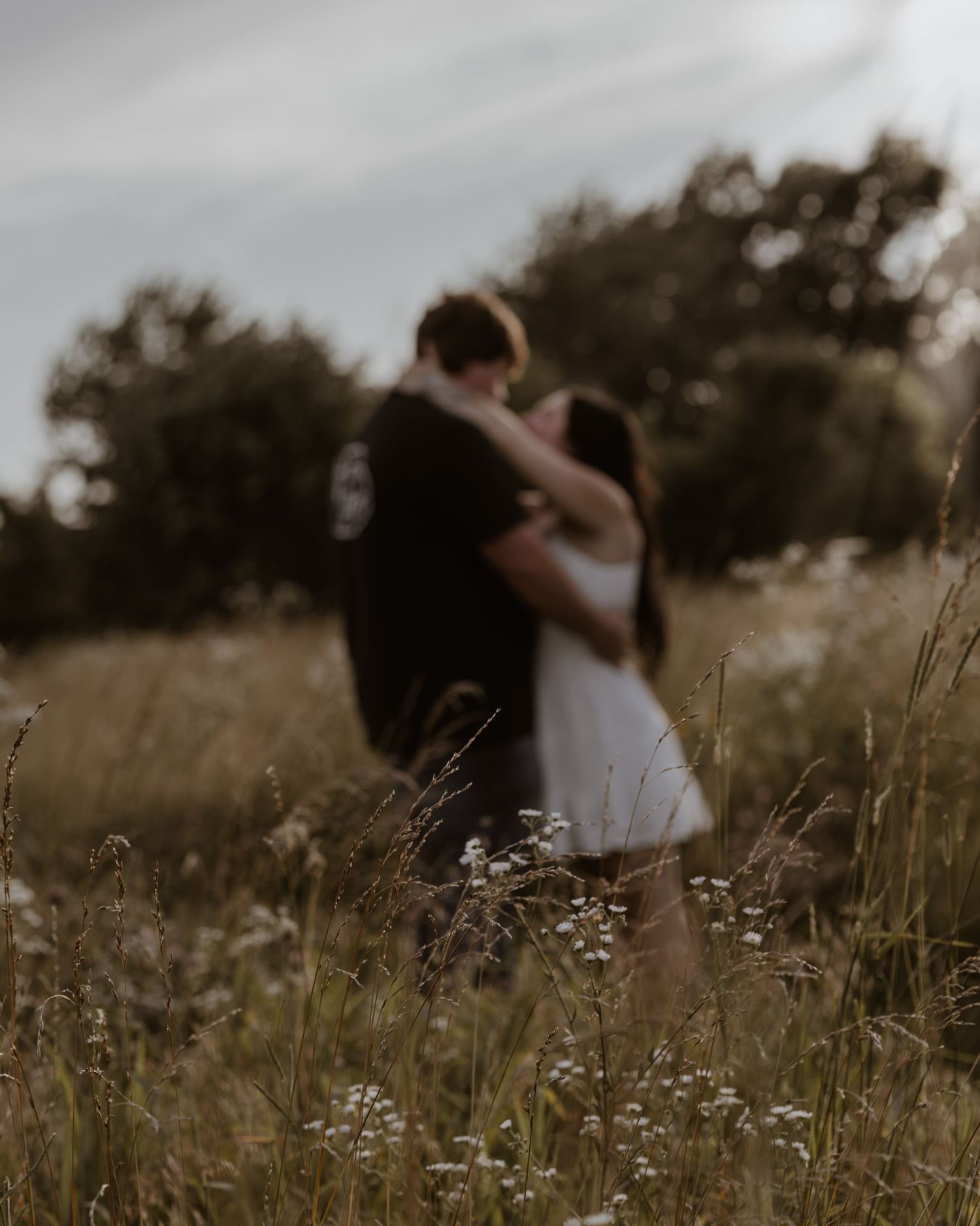 Couple embracing romantically in a wildflower meadow with tall grass and trees in soft bokeh background.