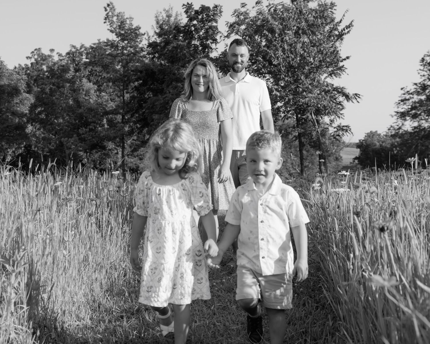 Black and white family photo of parents with two young children walking through tall grass in a field.