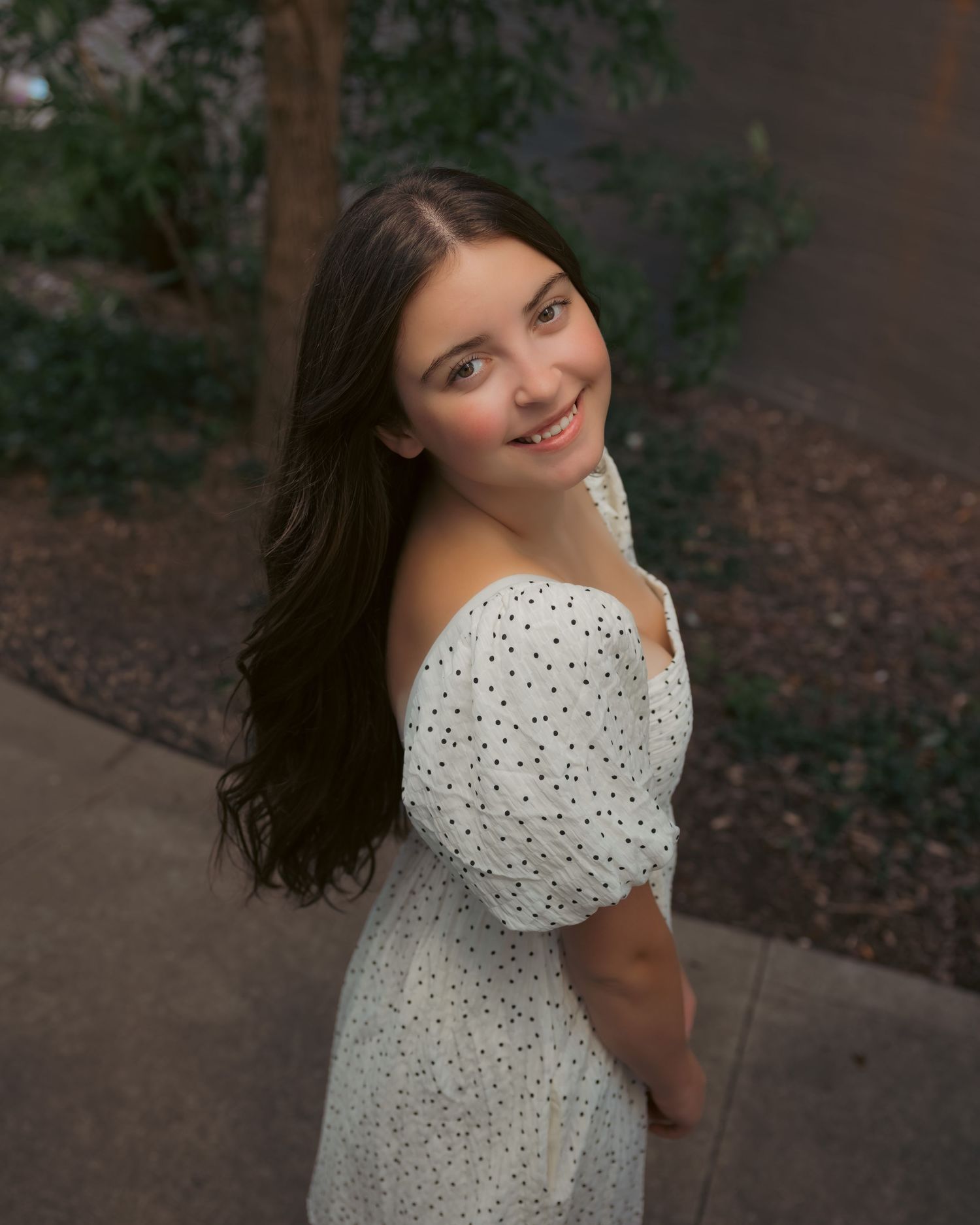 Young woman in white polka dot off-shoulder dress smiling over shoulder outdoors at dusk.