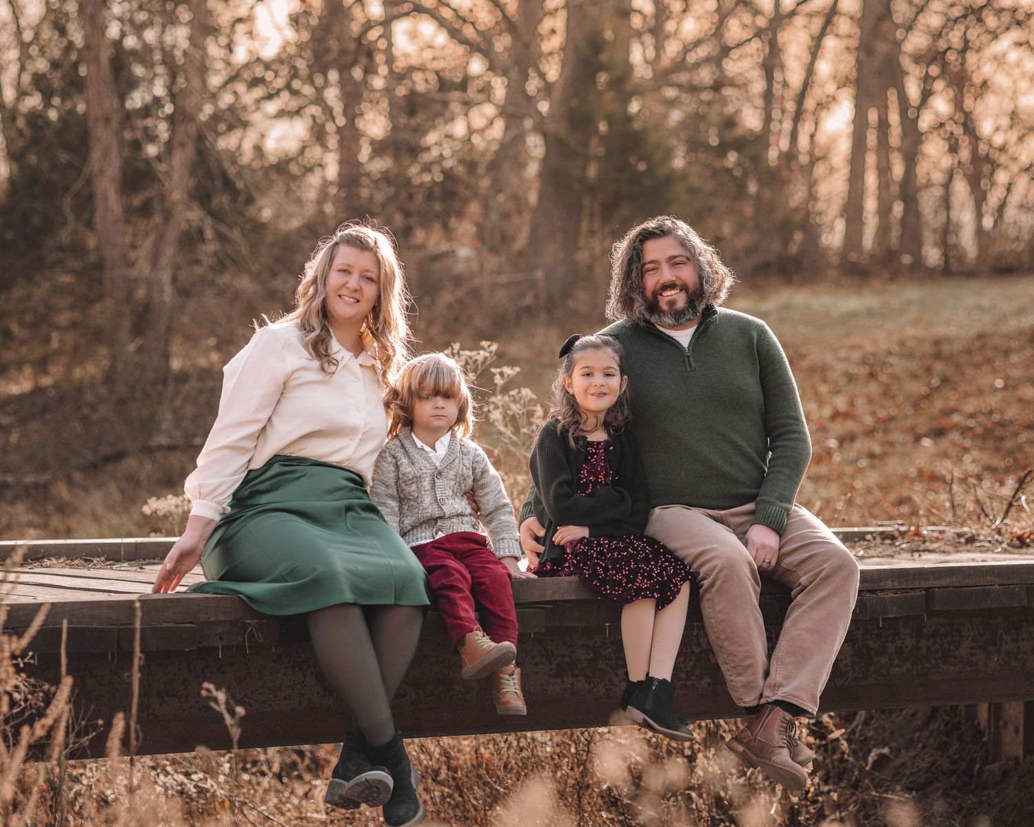 A family sits together on a rustic wooden fence during a warm autumn sunset in a wooded setting.