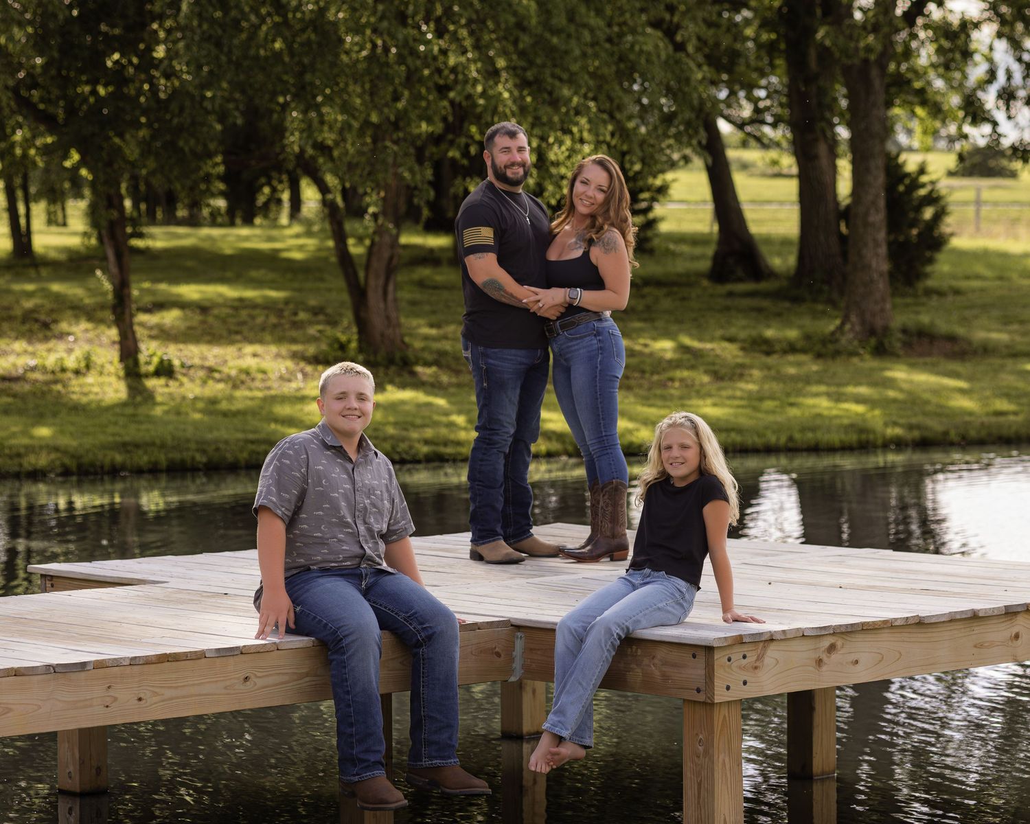 Family of four posing on a wooden dock by a pond surrounded by lush green trees on a sunny day.