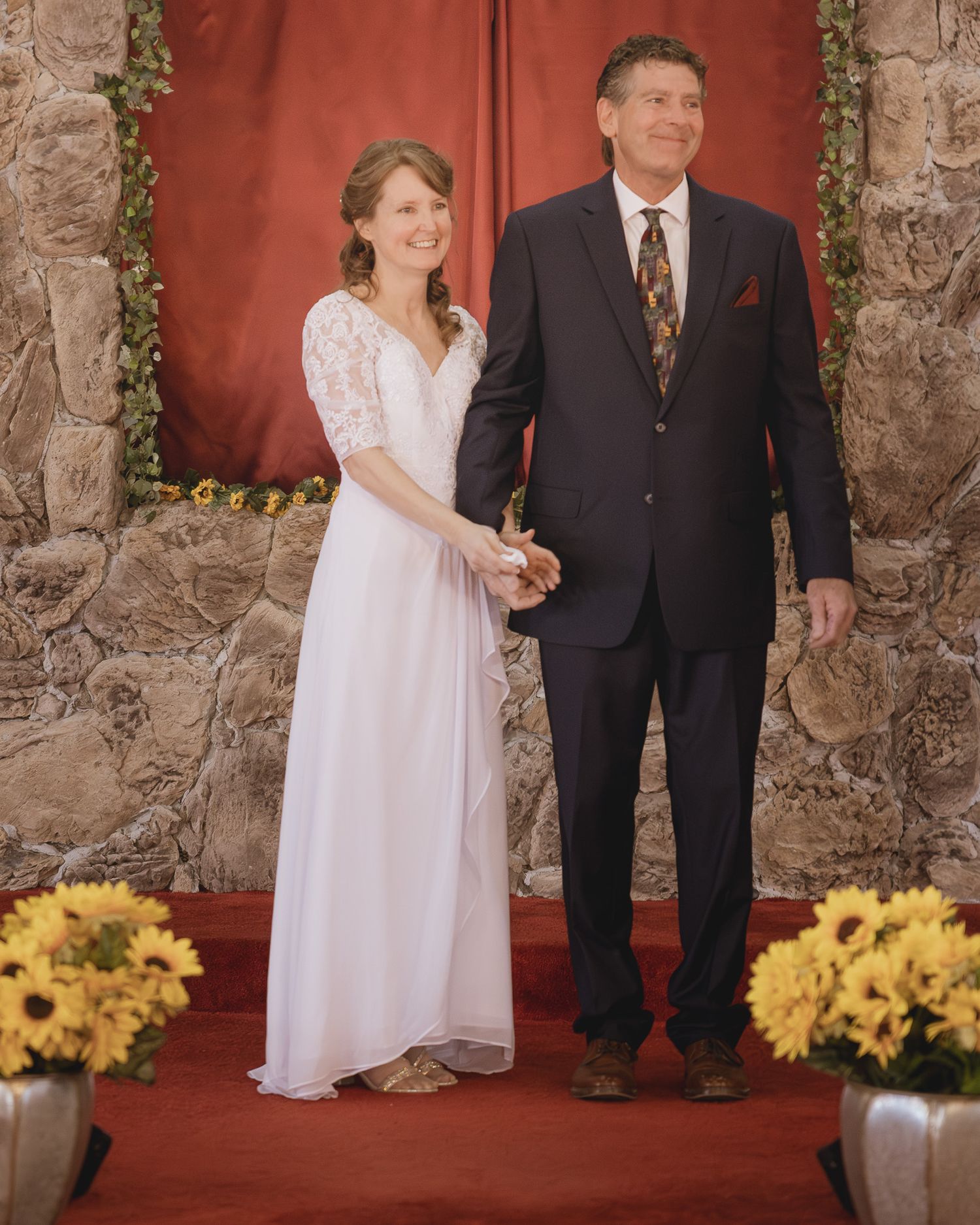 Couple in wedding attire holding hands at altar with sunflowers and stone wall backdrop.