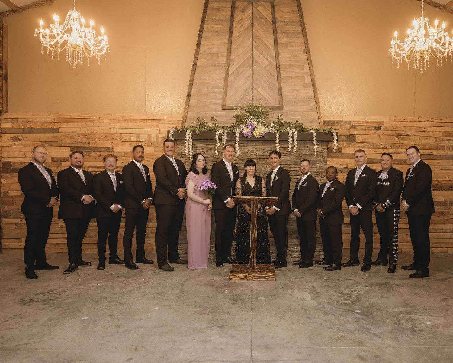 Wedding party poses in elegant barn venue with chandeliers, groomsmen in black suits flanking bride in lavender gown.