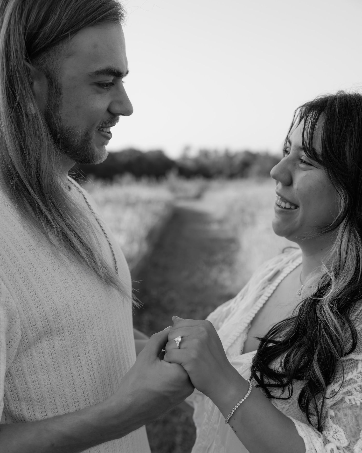 Black and white photo of a couple holding hands and smiling at each other in an outdoor field setting.