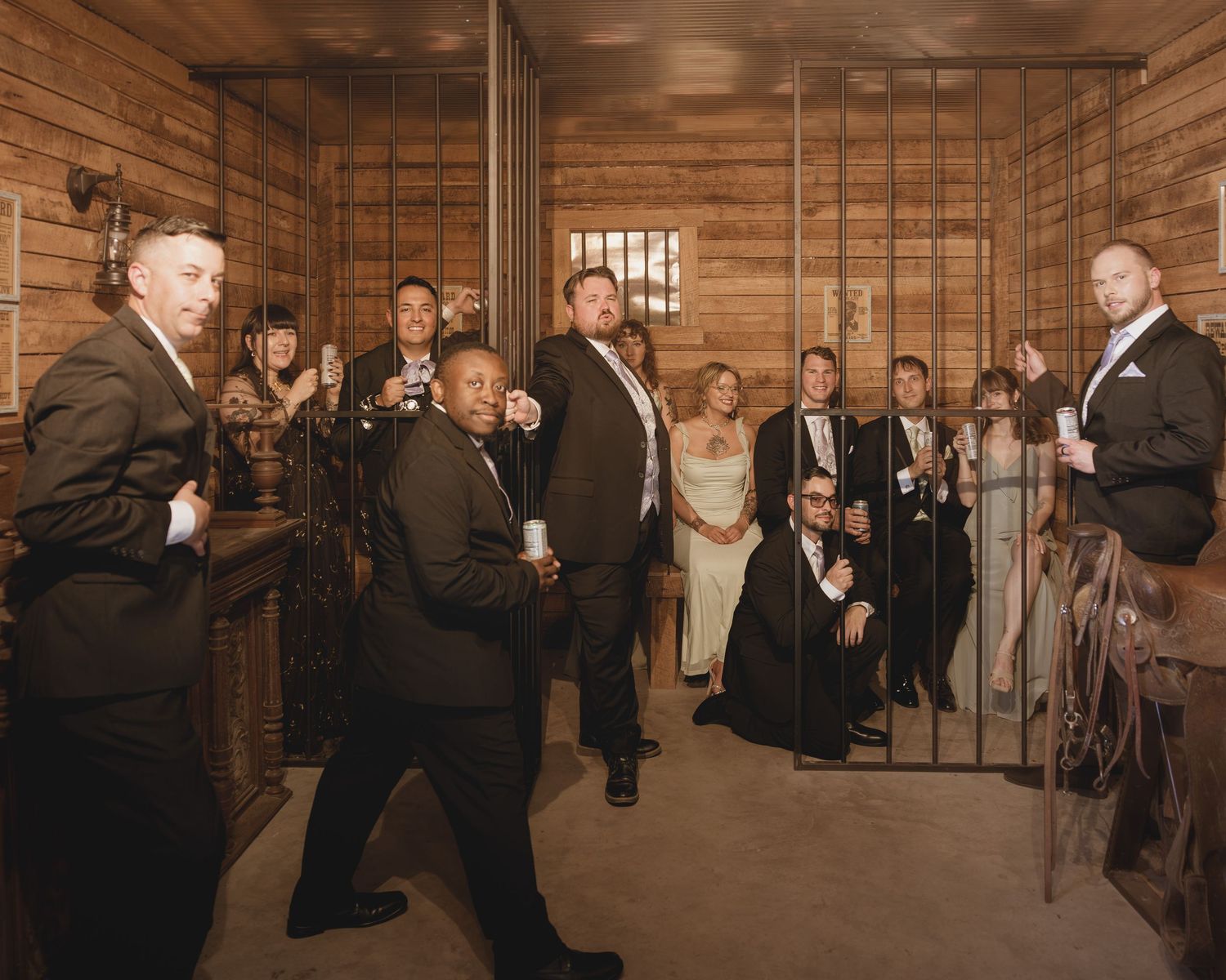 Formal group photo of wedding party posing in rustic wooden jail cell setting wearing elegant suits and gowns.