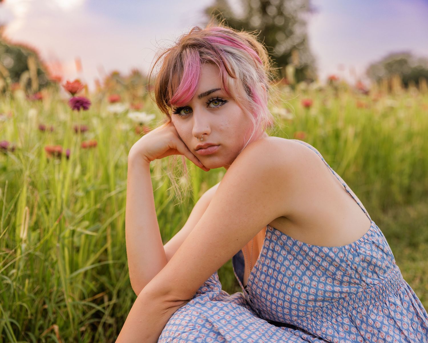 Young woman with pink hair sitting in a wildflower meadow wearing a blue sundress at golden hour.