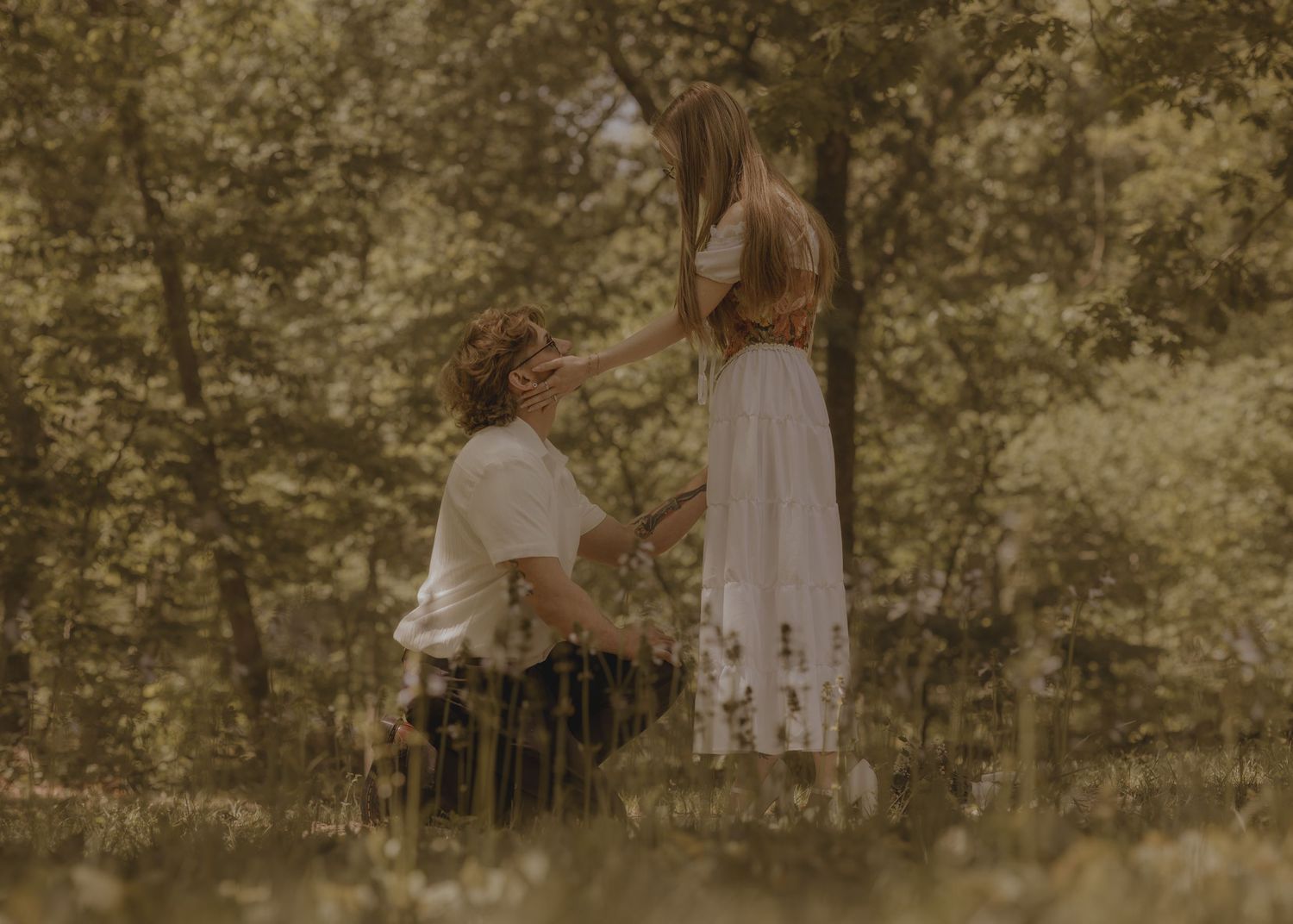 Person kneeling before partner in sunlit forest clearing, appearing to propose in romantic outdoor setting.