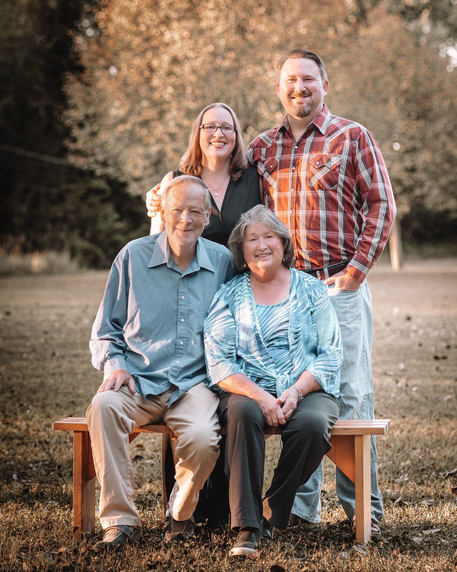 Family portrait of four adults posing outdoors, two seated on a bench and two standing behind them.