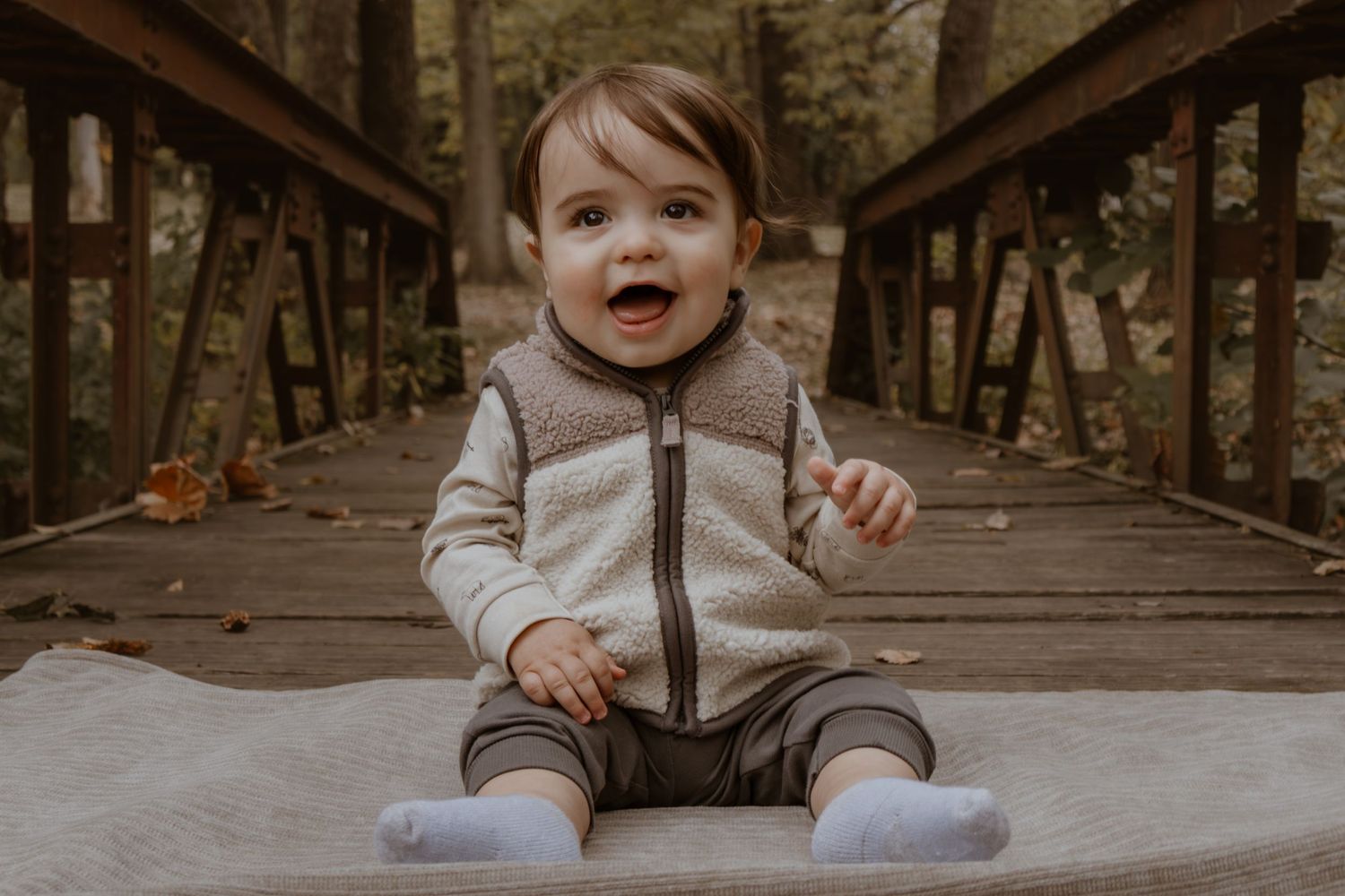 A toddler in a zip-up sweater sits smiling on a wooden bridge.