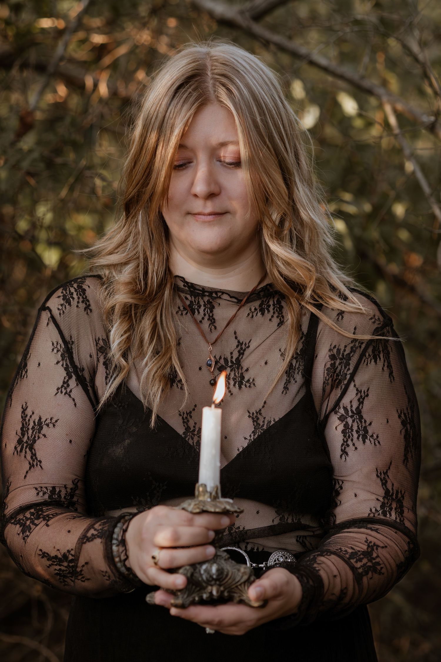 Figure in black lace dress holding a lit candle in a moody forest setting.