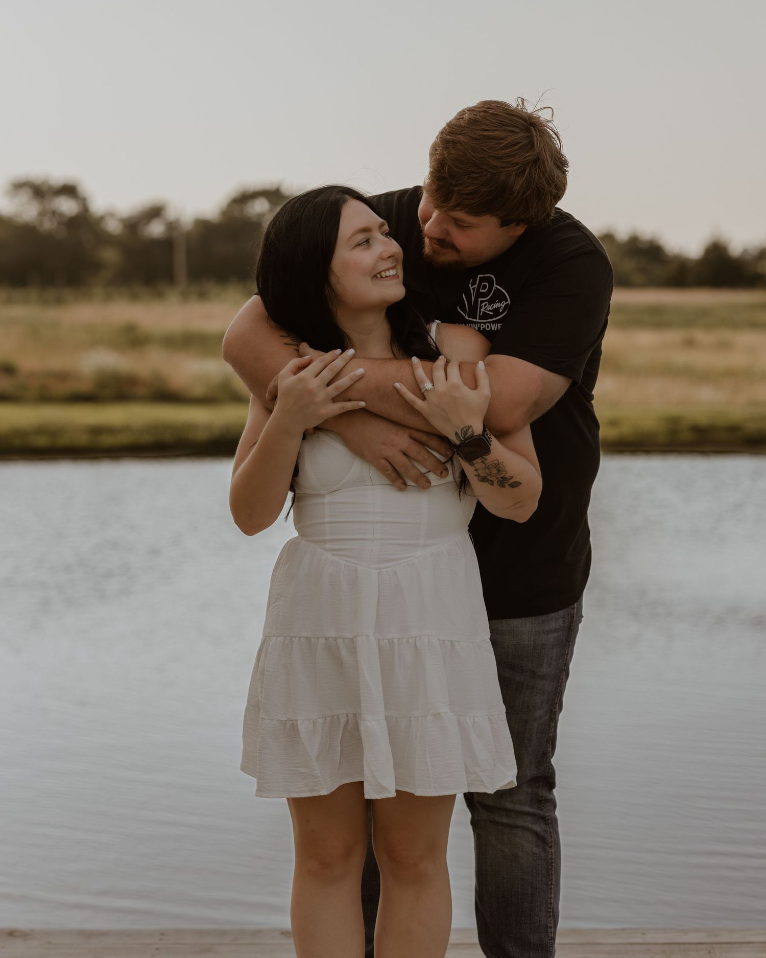 Couple embracing by a lake at sunset, woman in white dress smiling as man holds her from behind.