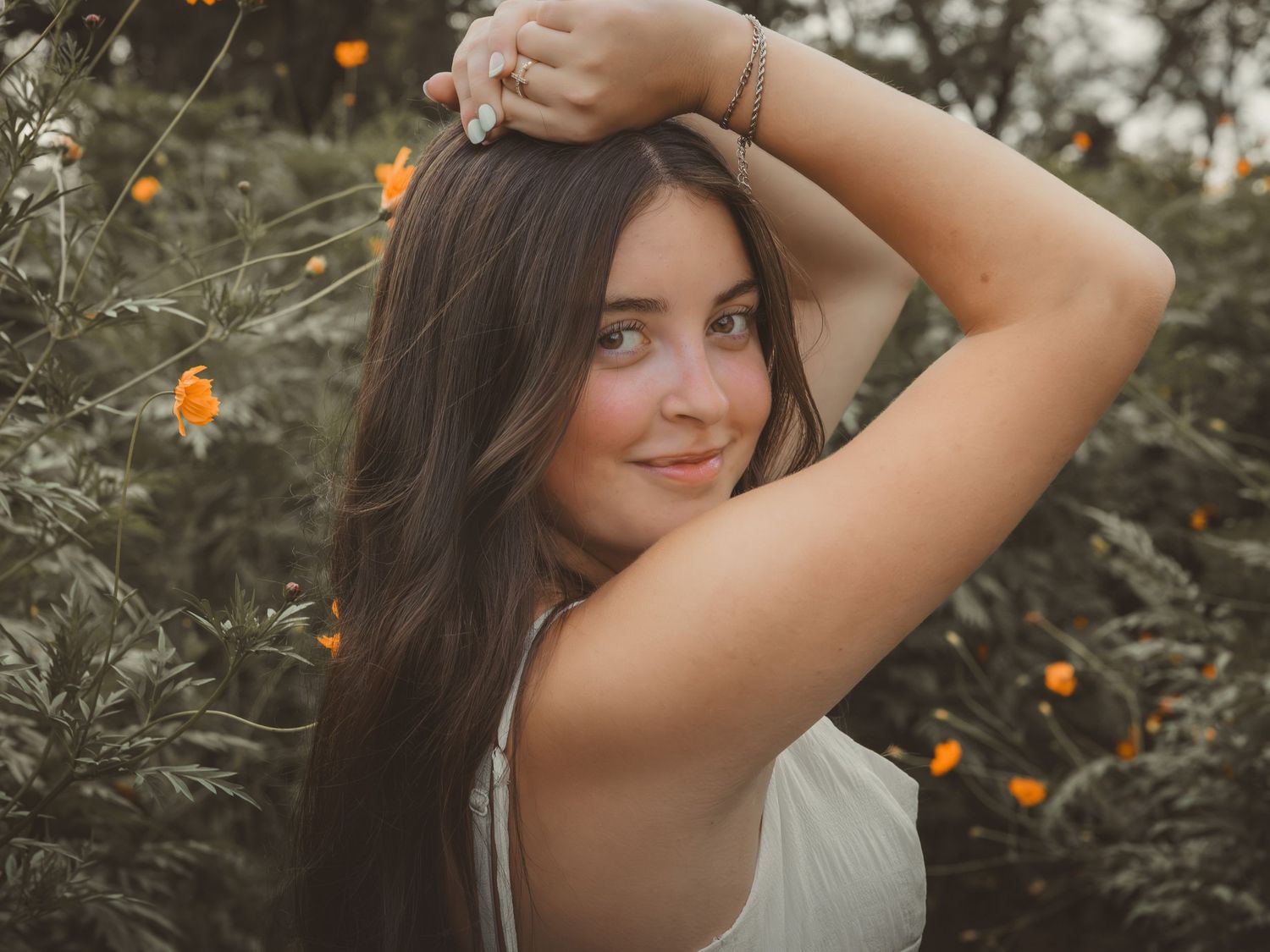 Young woman with long dark hair poses outdoors among orange flowers, wearing a white top in soft natural light.