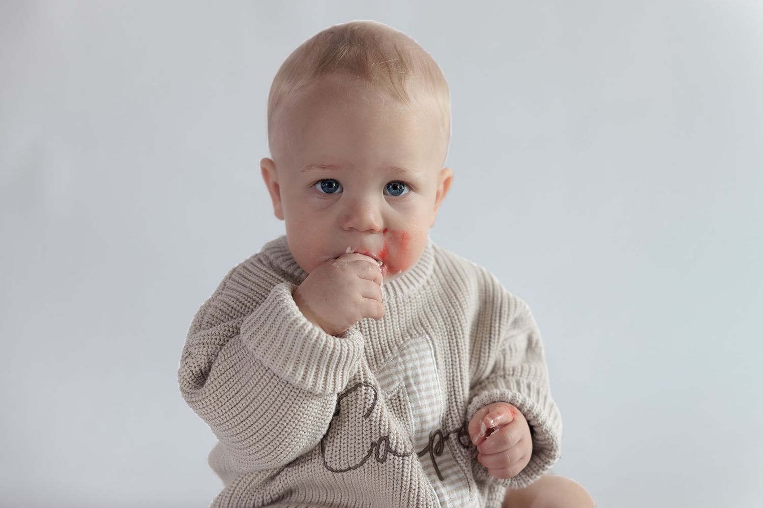 Adorable baby in a beige knit sweater with finger in mouth, sitting against a white background.