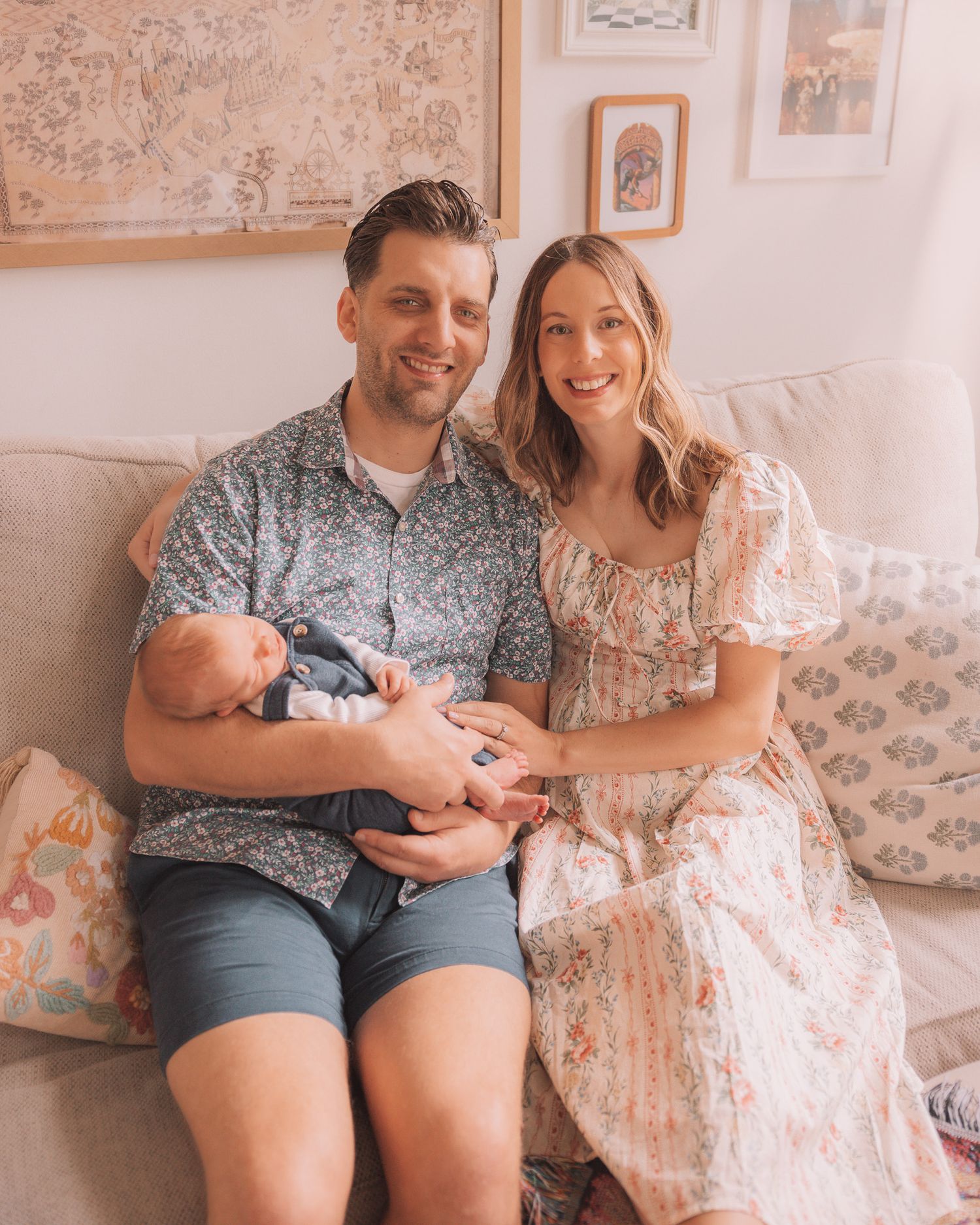 Smiling couple with newborn baby sits together on a light couch at home.