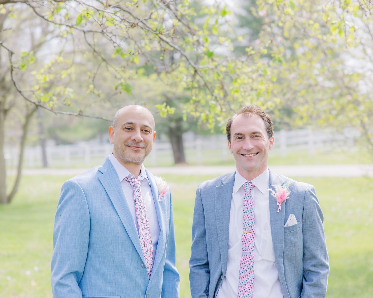 Two grooms in light blue suits with pink ties and boutonnieres smiling outdoors at a spring wedding.
