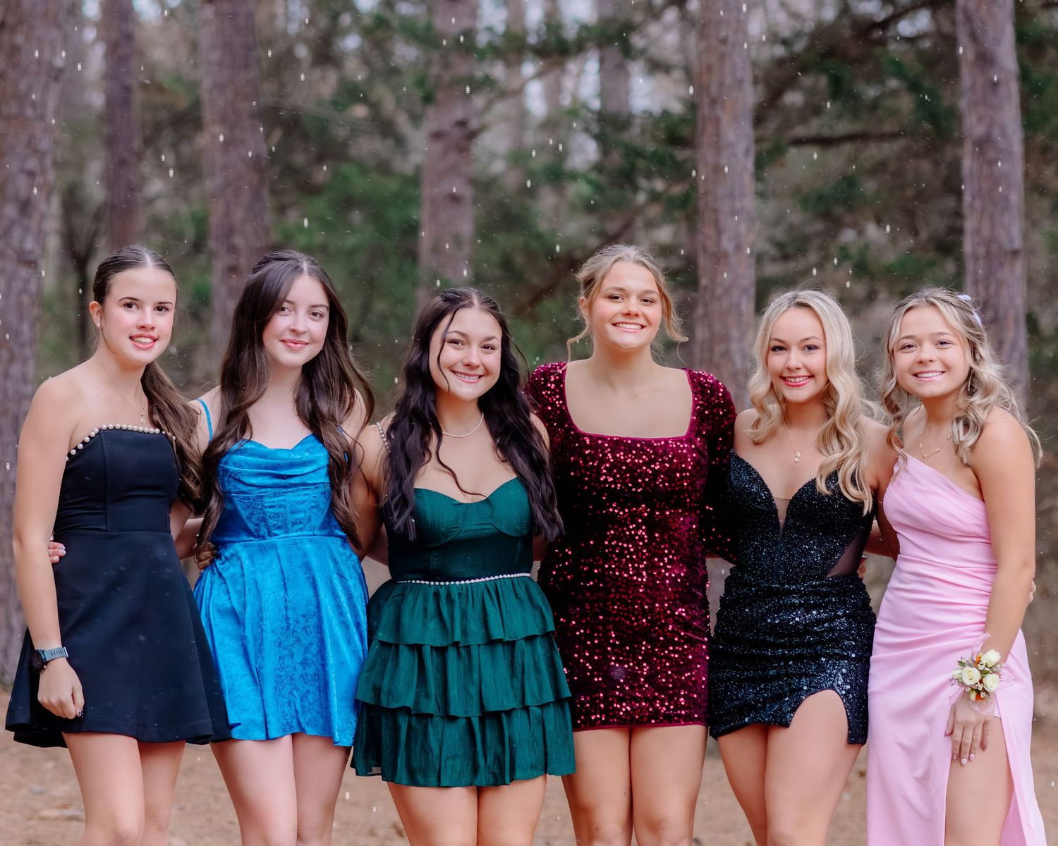 A group of young women in formal dresses stand together outdoors in a wooded setting for a special occasion photo.