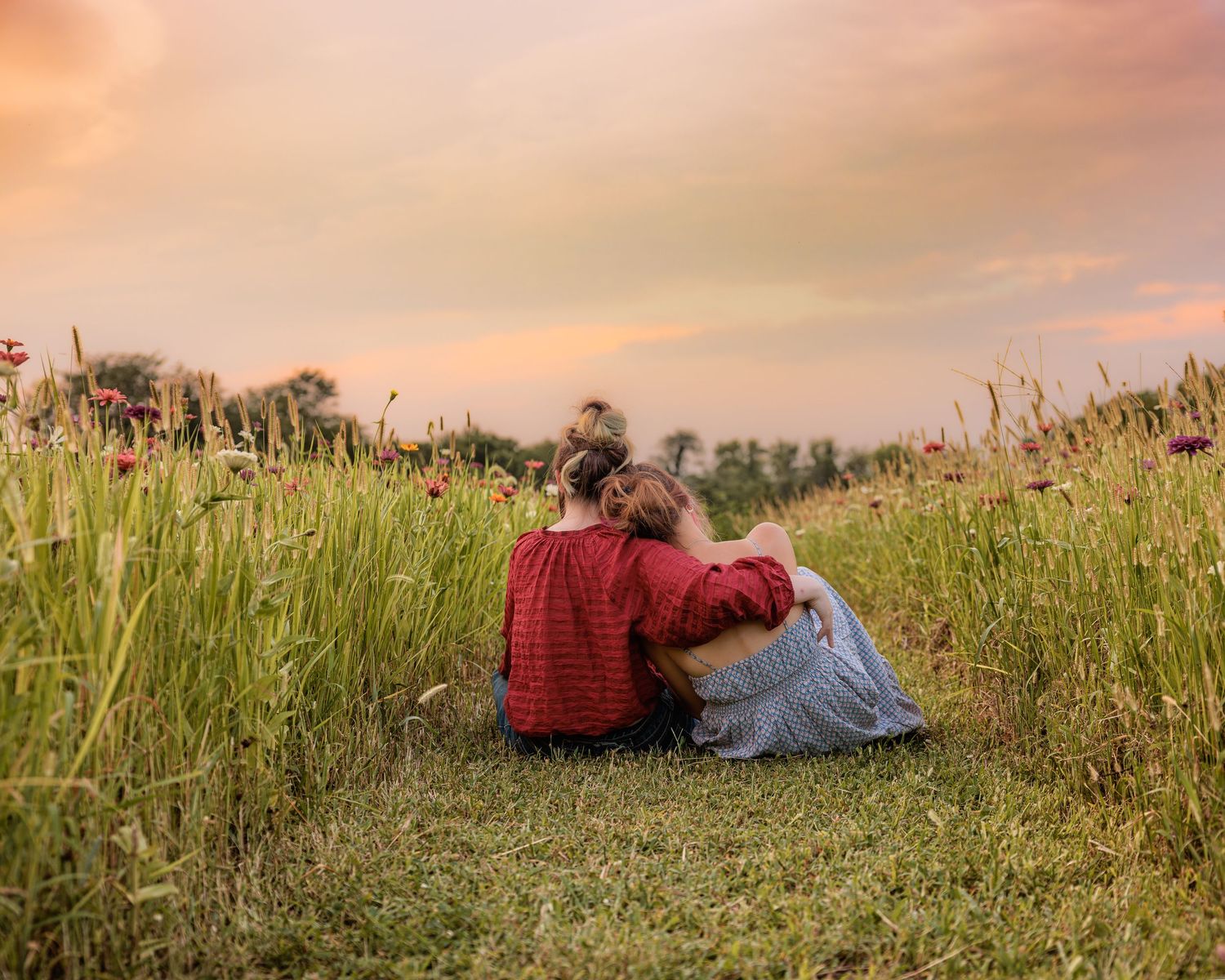 Two people sitting together in a wildflower field at sunset, sharing a tender embrace amid tall grass.