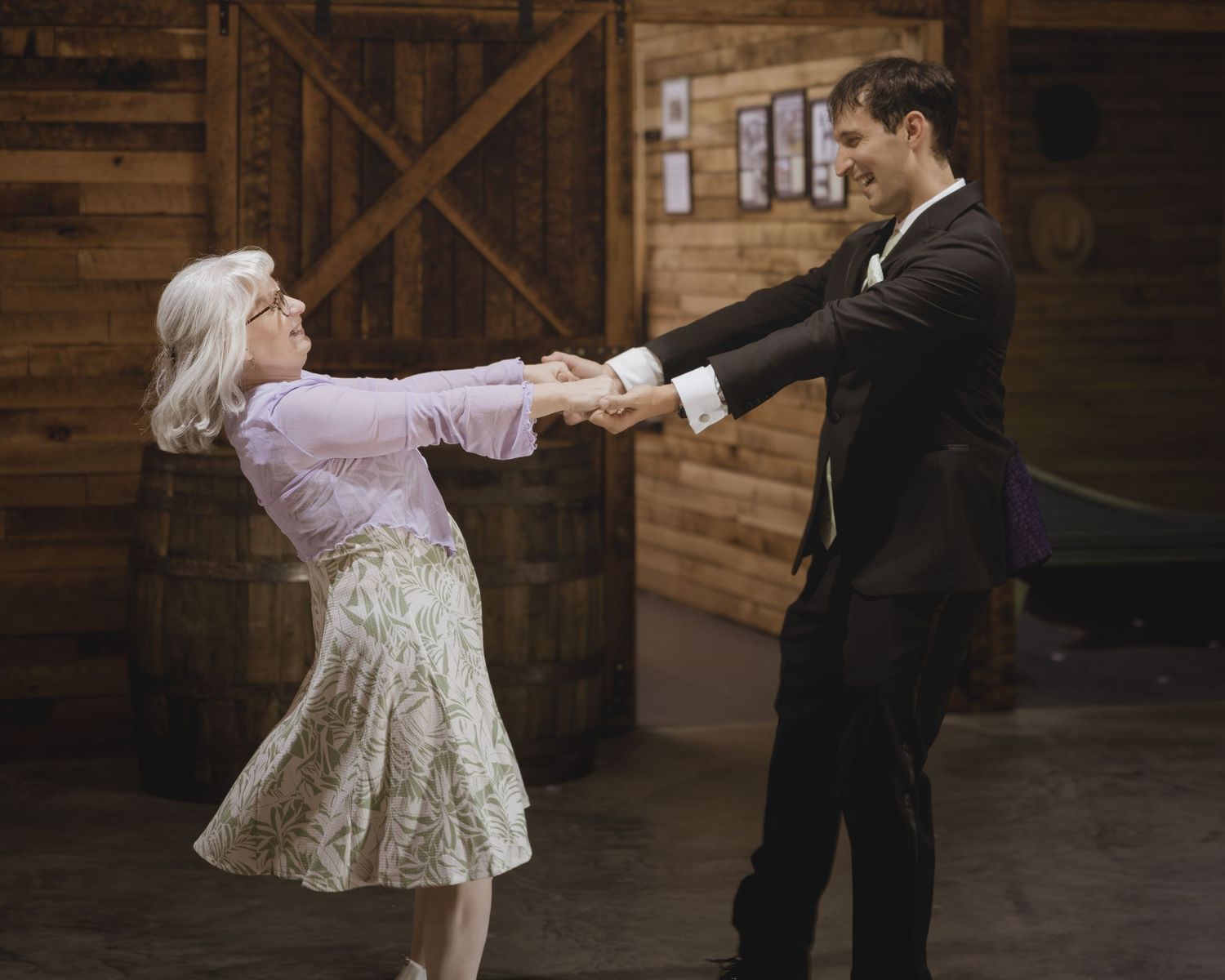 Elderly woman and young man in formal attire dancing joyfully together in a rustic barn setting.