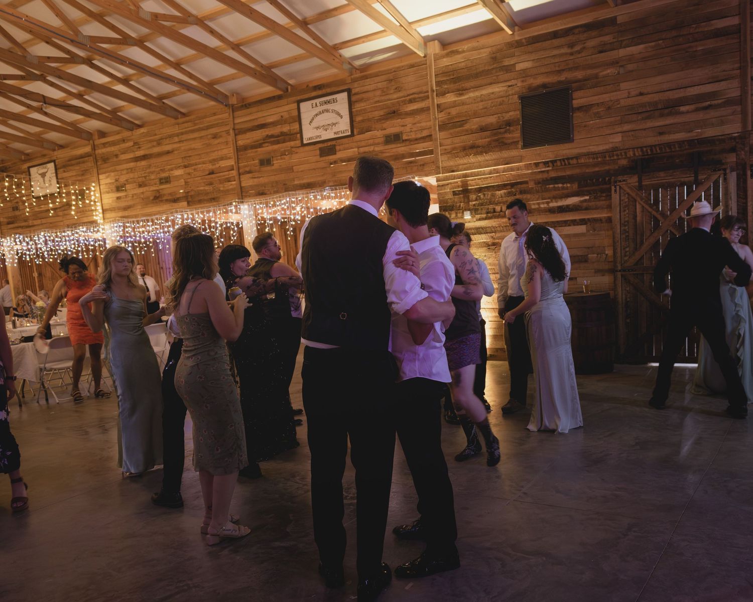 Guests dancing together on the floor of a warmly lit rustic barn wedding reception with string lights.
