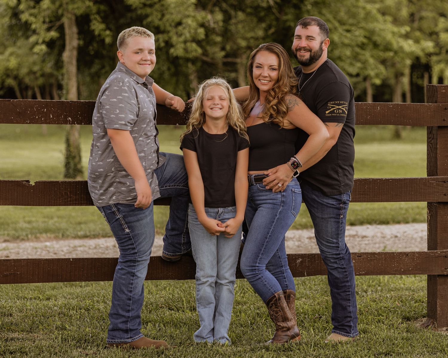 Family of four posing and smiling together by a rustic wooden fence outdoors.