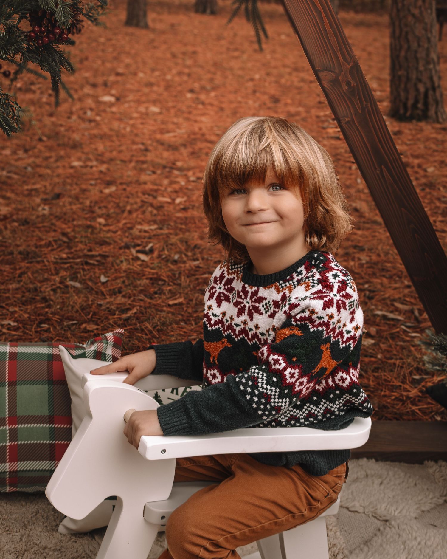 Child wearing a festive holiday sweater sits on a white chair against an autumn backdrop with red-orange ground.