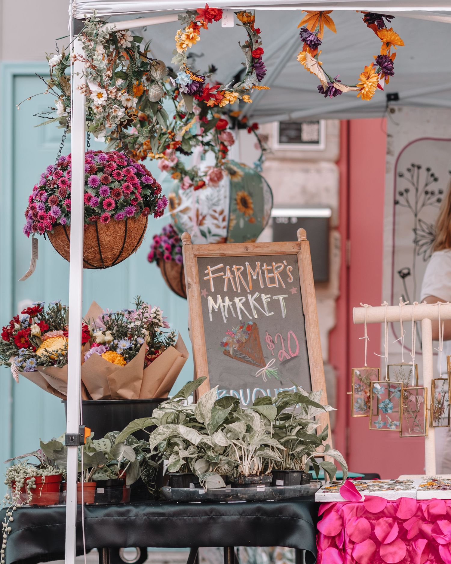 Colorful hanging flower baskets and potted plants decorate a farmers market display with pink and rustic signage.