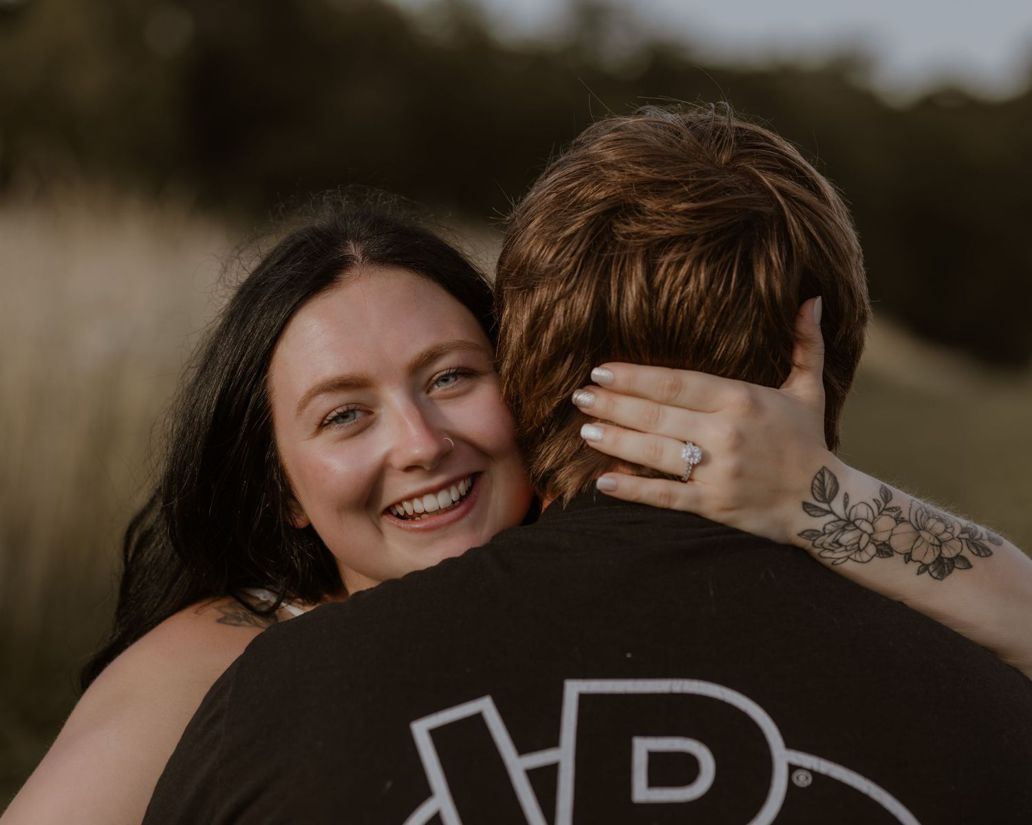 Joyful woman with tattoos hugs her partner tightly, smiling broadly during outdoor engagement shoot.