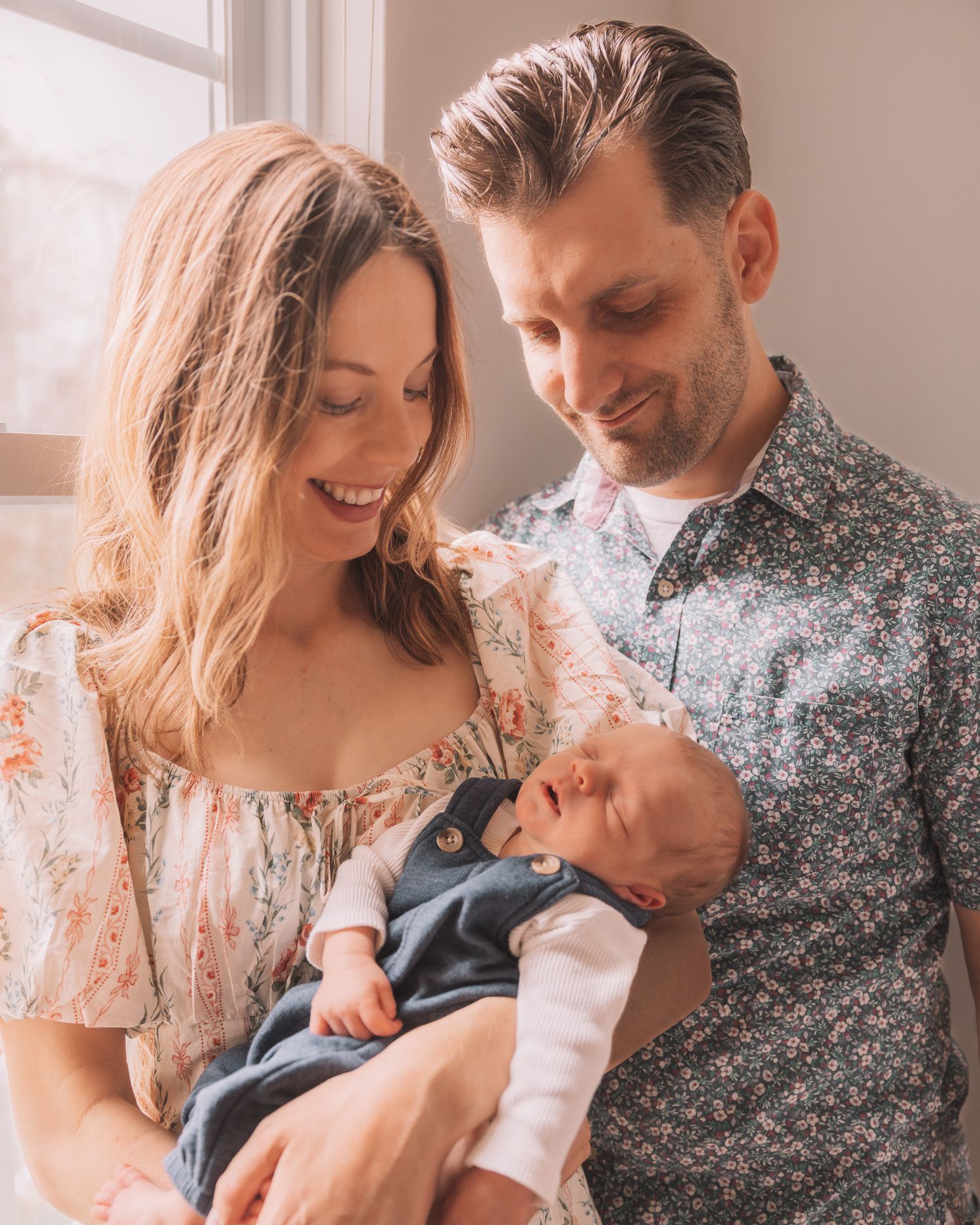 Smiling couple in floral outfits lovingly gazing at their newborn baby indoors near a bright window.