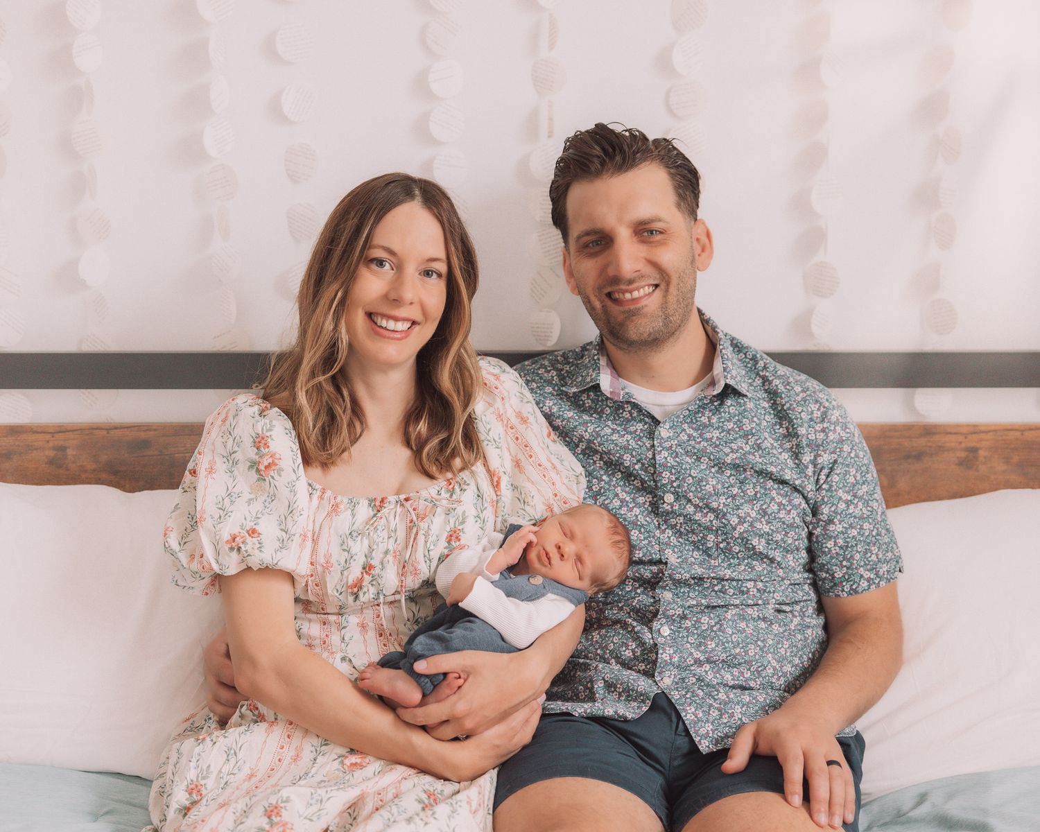 Smiling couple sitting on a bed holding their newborn baby, woman in floral dress and man in blue floral shirt.