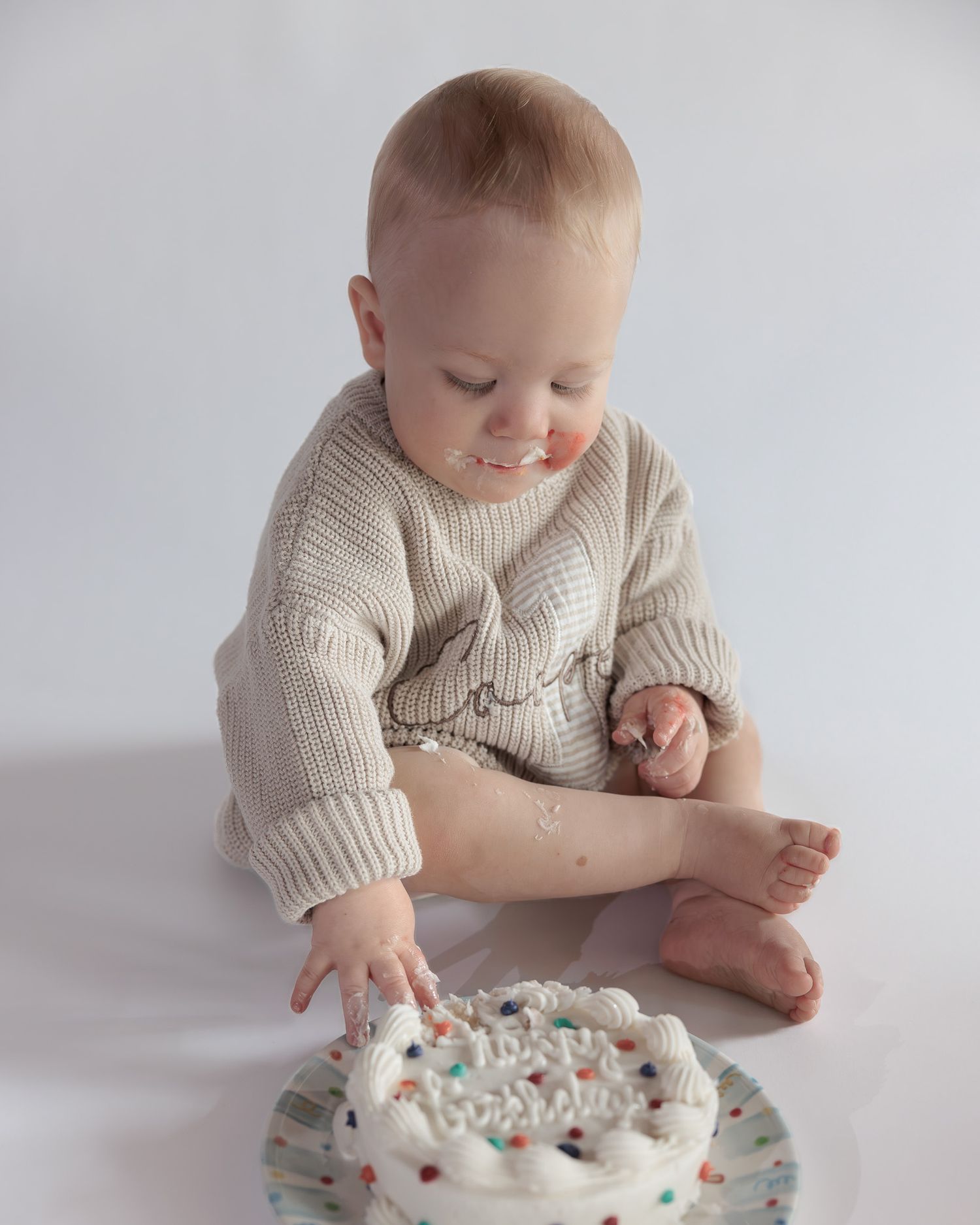 Smiling baby in beige knit romper reaches into white frosted smash cake with colorful sprinkles.