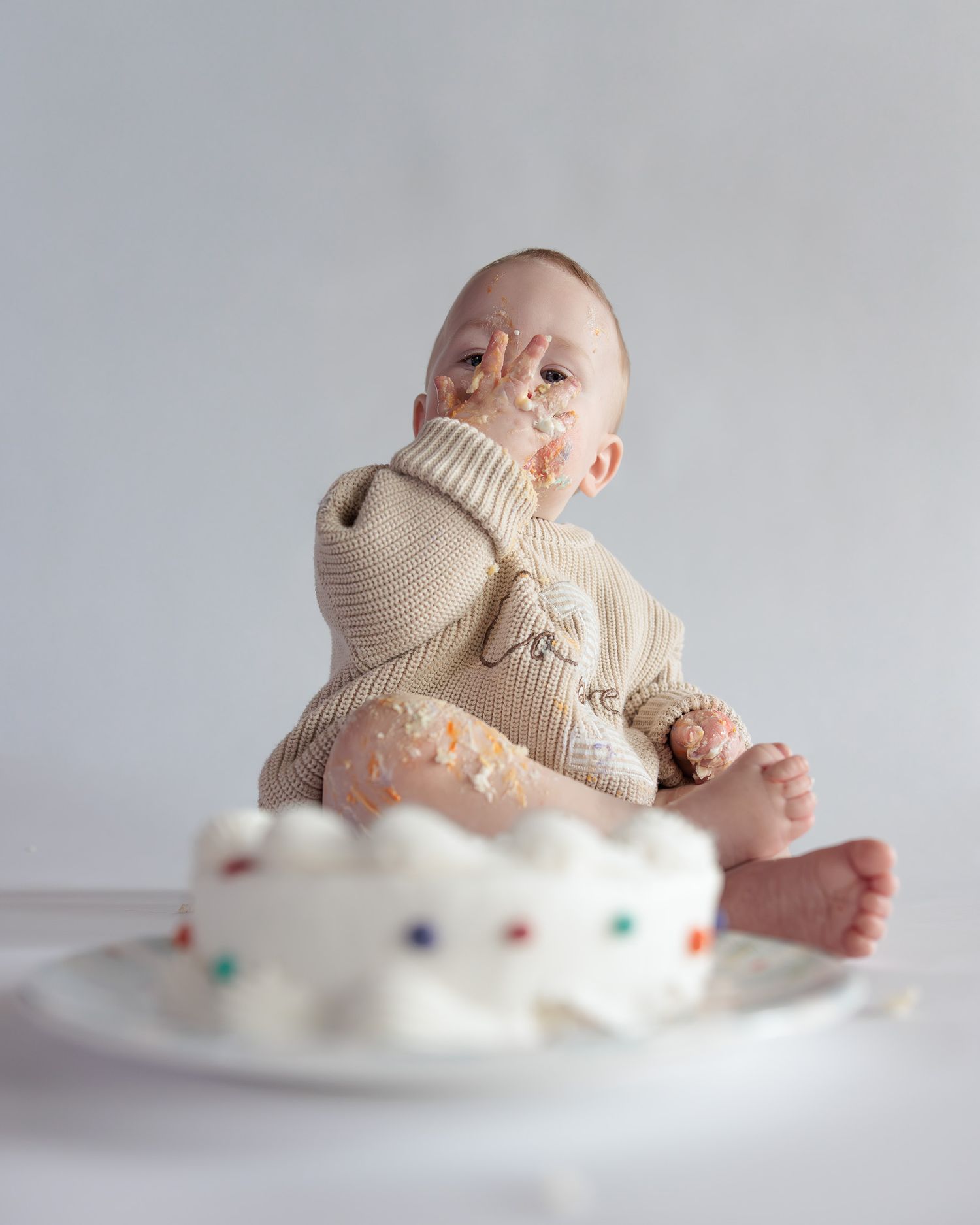 Messy baby boy covered in white frosting during first birthday cake smash session wearing beige knit romper on white background.