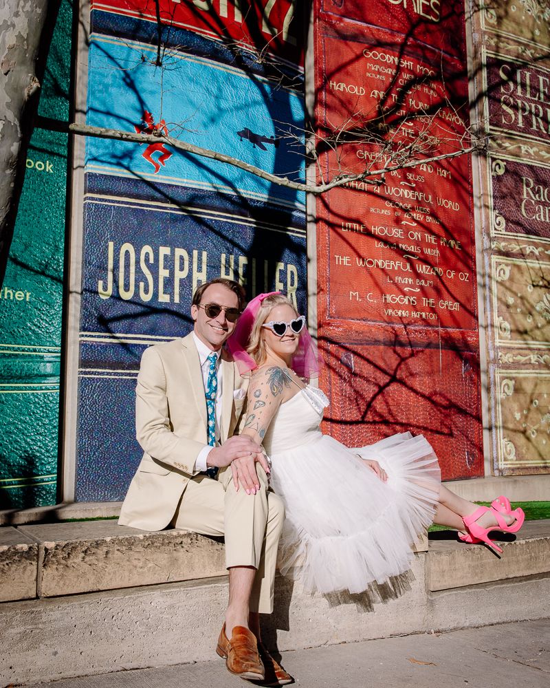 A couple in stylish attire poses against a colorful wall with blue and red sections near a door marked 'Joseph'.