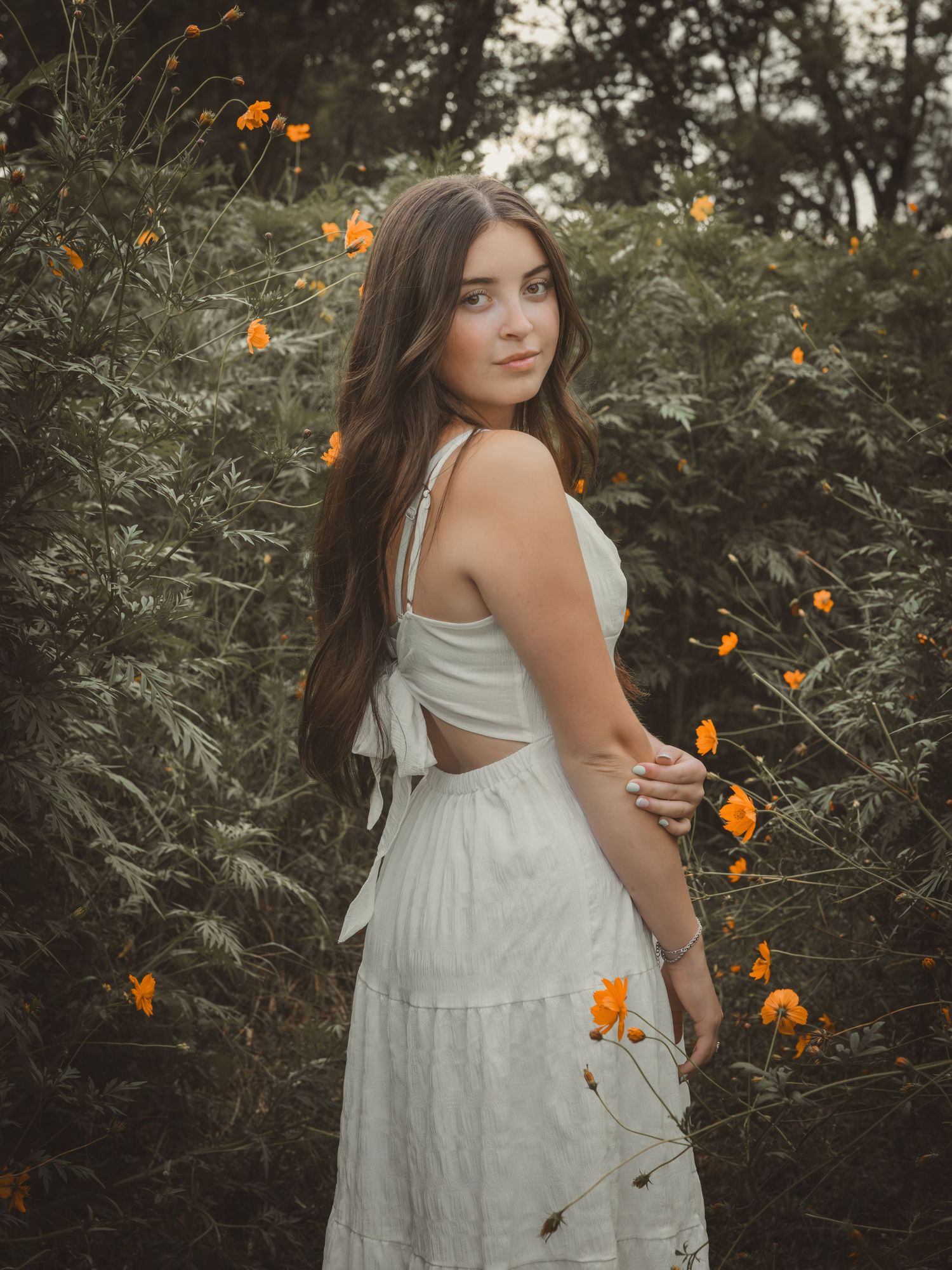 Young woman in white dress poses among orange wildflowers and green foliage in a dreamy outdoor portrait.