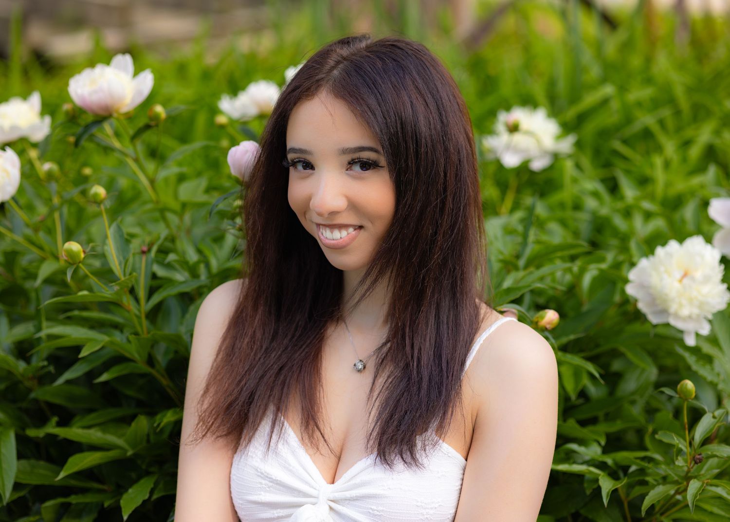 Young woman with long dark hair smiling outdoors among blooming white peony flowers in a garden.