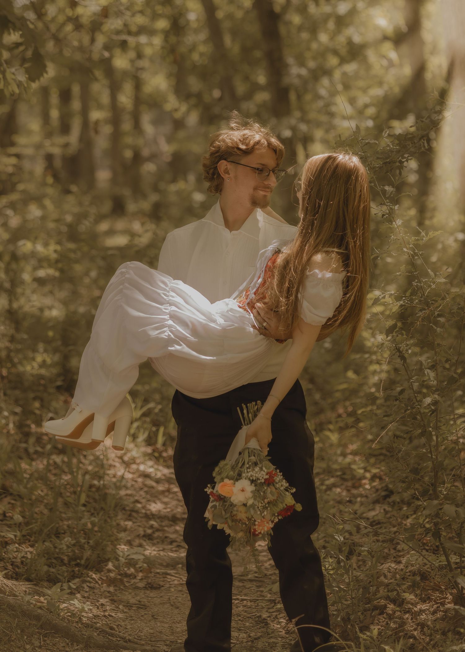Groom carrying bride in white dress through a sunlit forest, holding a floral bouquet in a romantic wedding photo.