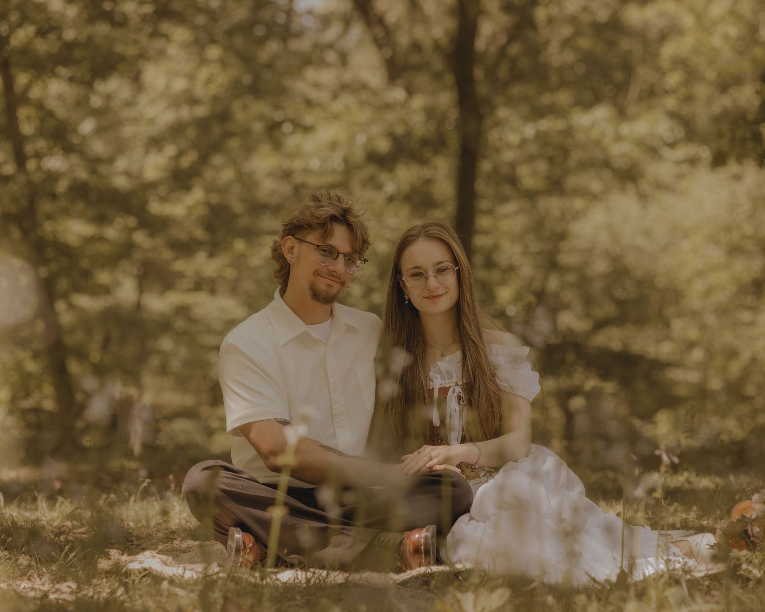 Couple sitting together on grass in a sunlit forest during an outdoor engagement photo session.