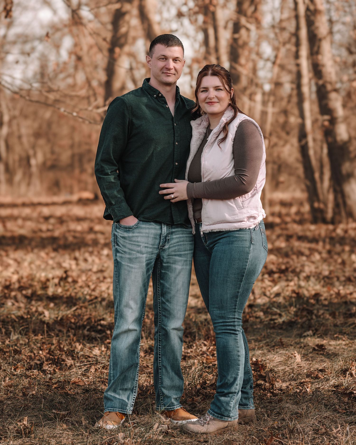 Couple in casual attire embracing while standing in a wooded area with autumn foliage.