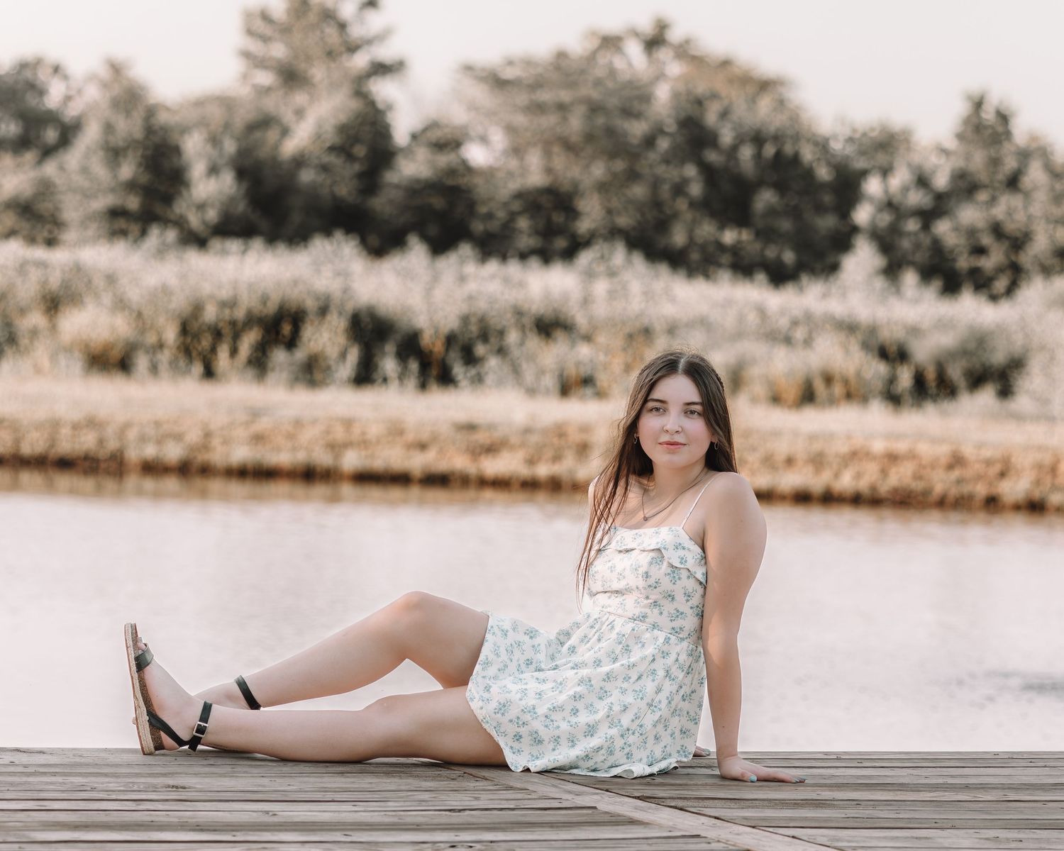 A woman in a floral dress poses on a wooden dock by a serene lake.