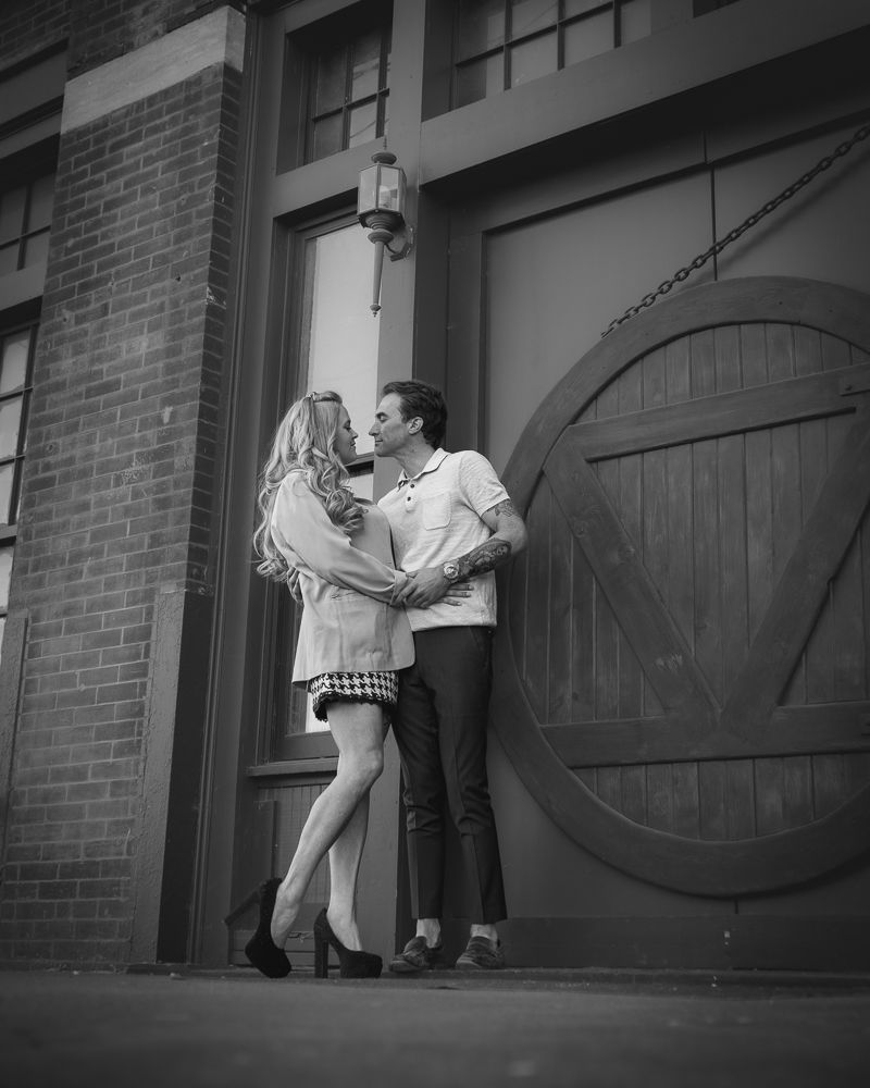 A romantic couple embraces by a rustic wooden door on a brick building in black and white.