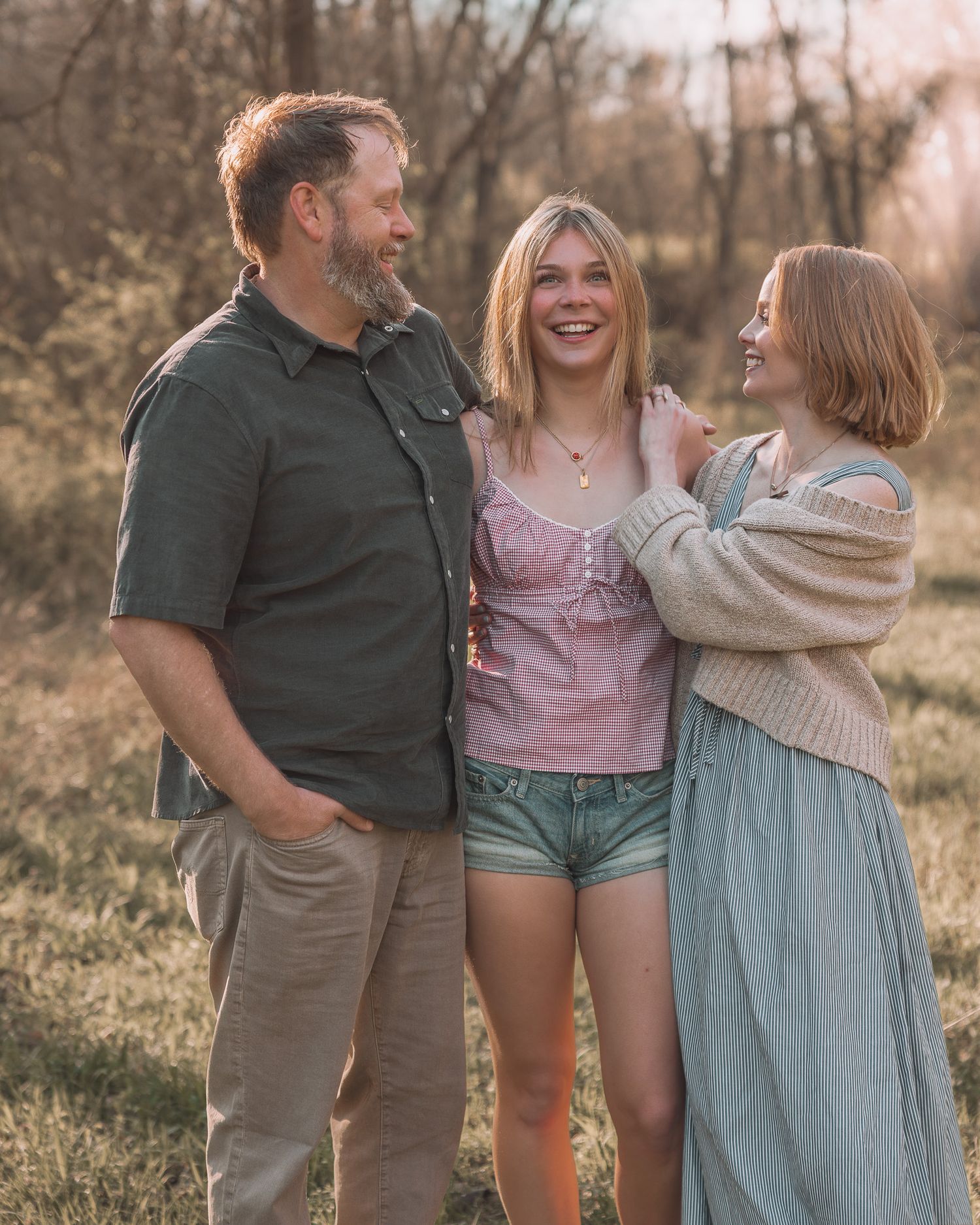 Happy family of three laughing together in a sunlit outdoor field setting.