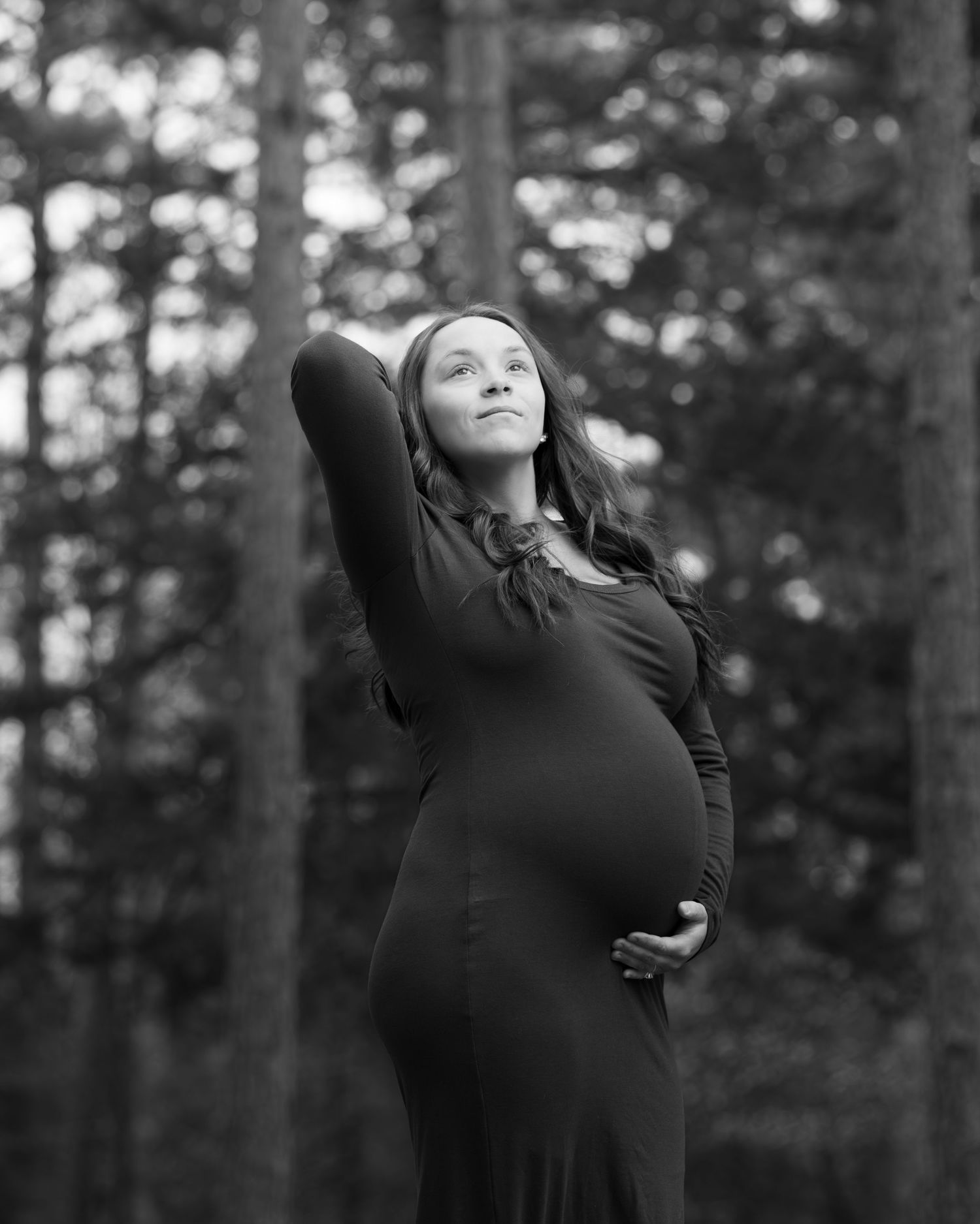 Pregnant woman in black dress cradling her baby bump outdoors among trees in a serene black and white maternity photo.