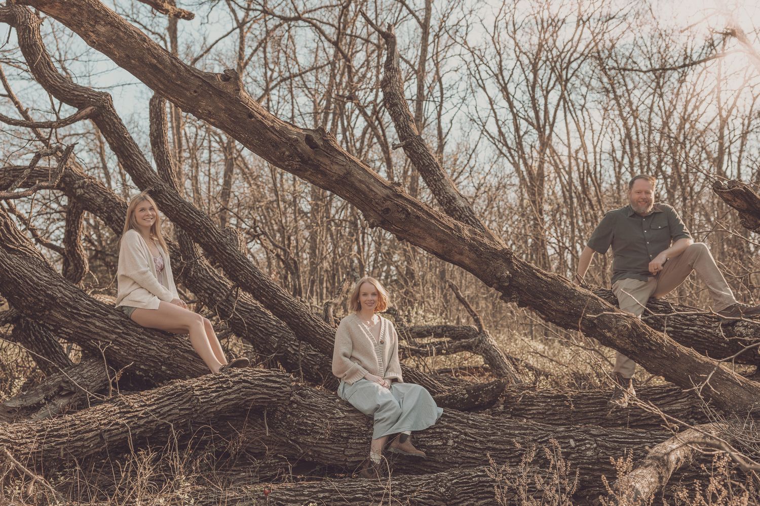 Three people sitting on fallen tree branches in a bare winter forest during golden hour.