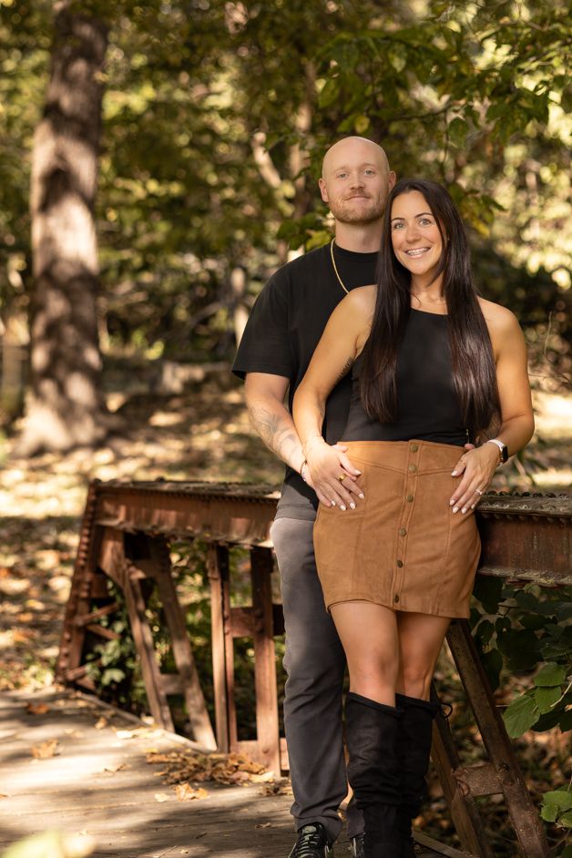 Couple posing on a rustic bridge in a forest, woman in brown skirt and black top, man in black shirt and gray pants.