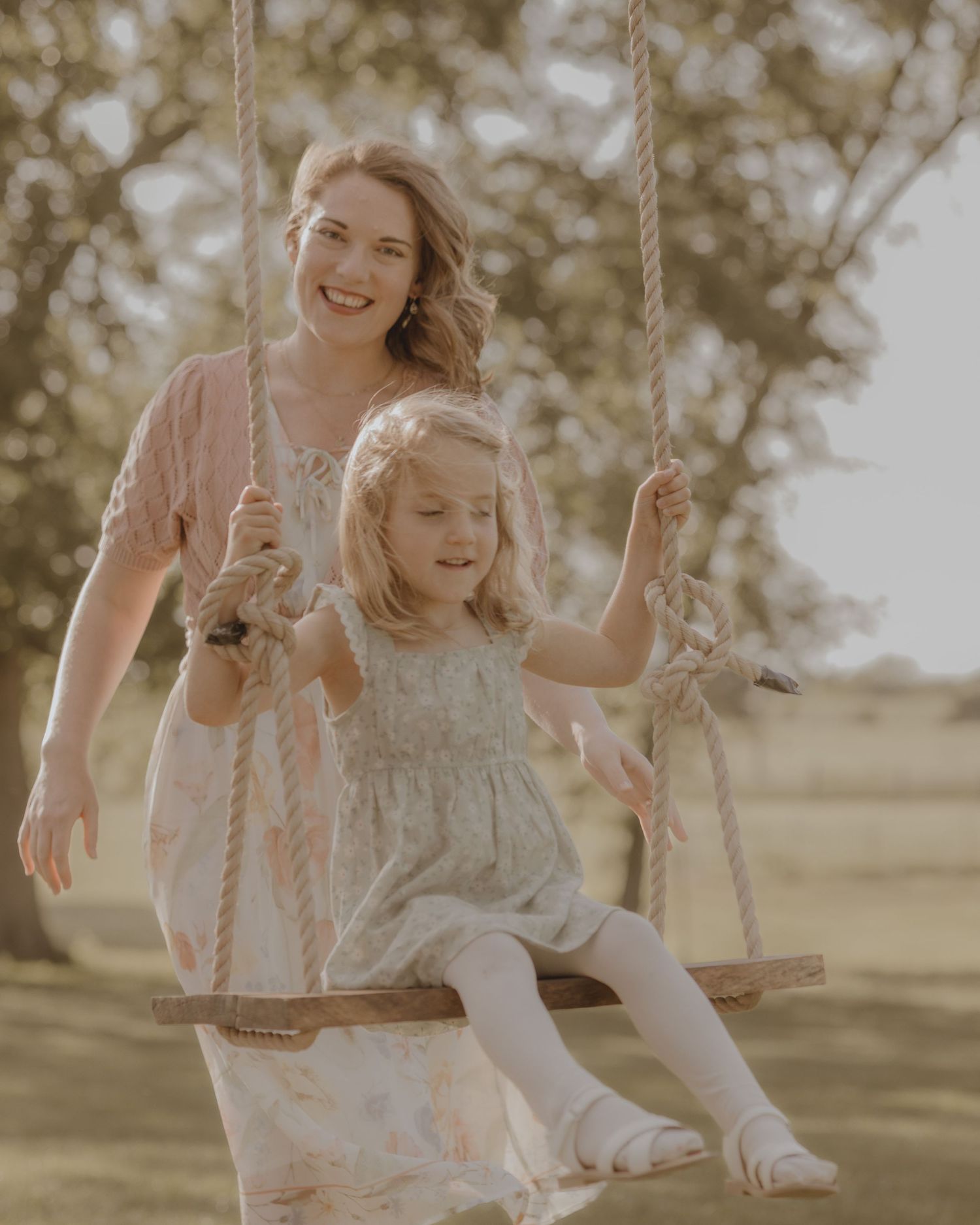 A smiling mother pushes her young blonde daughter on a wooden rope swing outdoors.