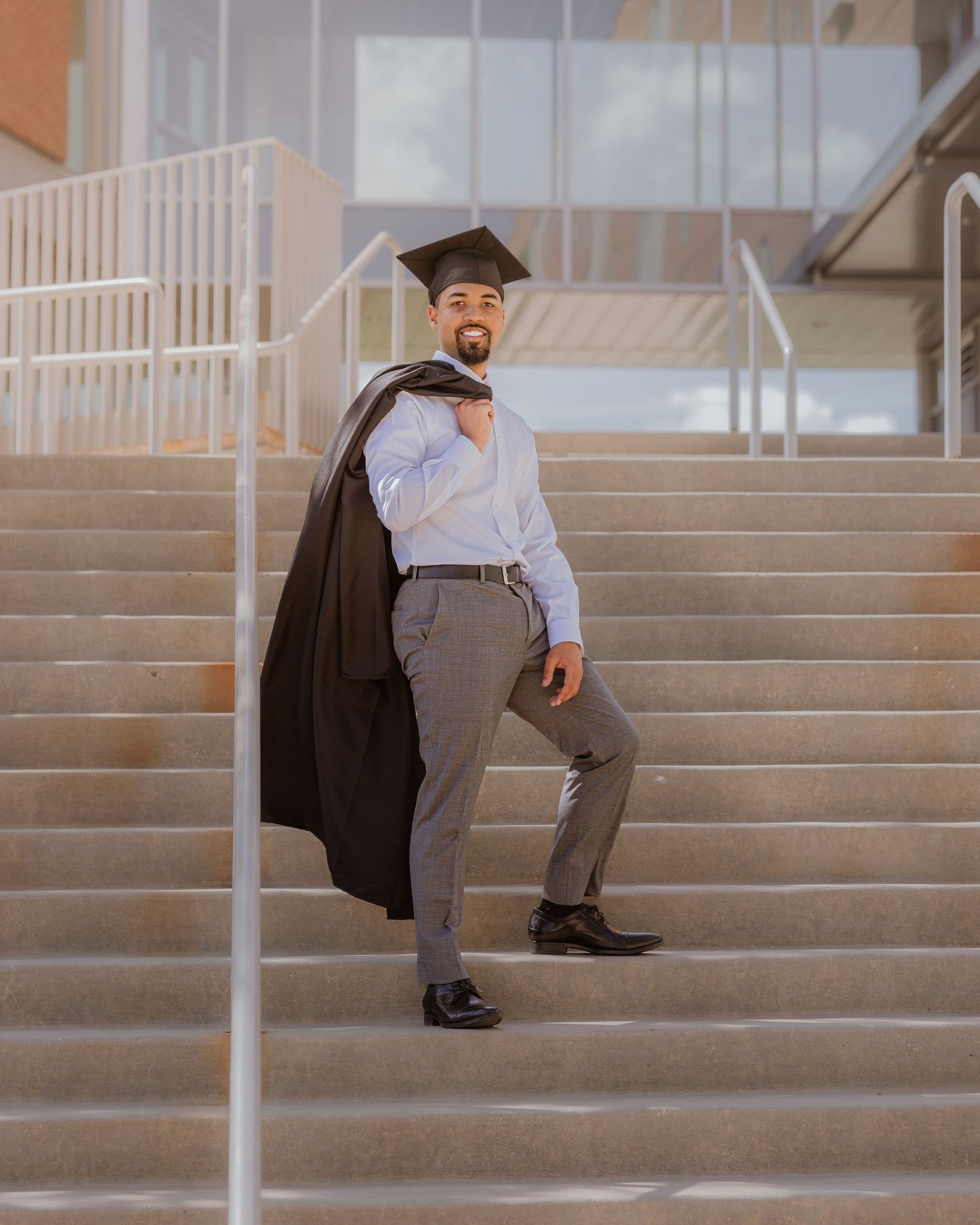 Graduate poses on steps holding black gown over shoulder, wearing cap and dress clothes.