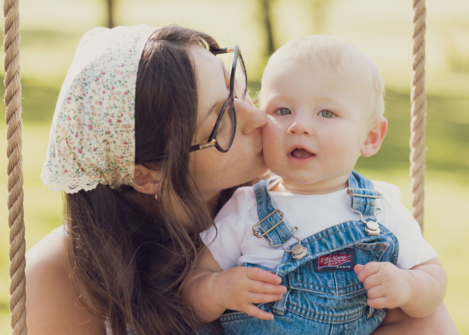 A mother with glasses kisses her baby boy on a swing, dressed in denim overalls, in a sunny outdoor setting.