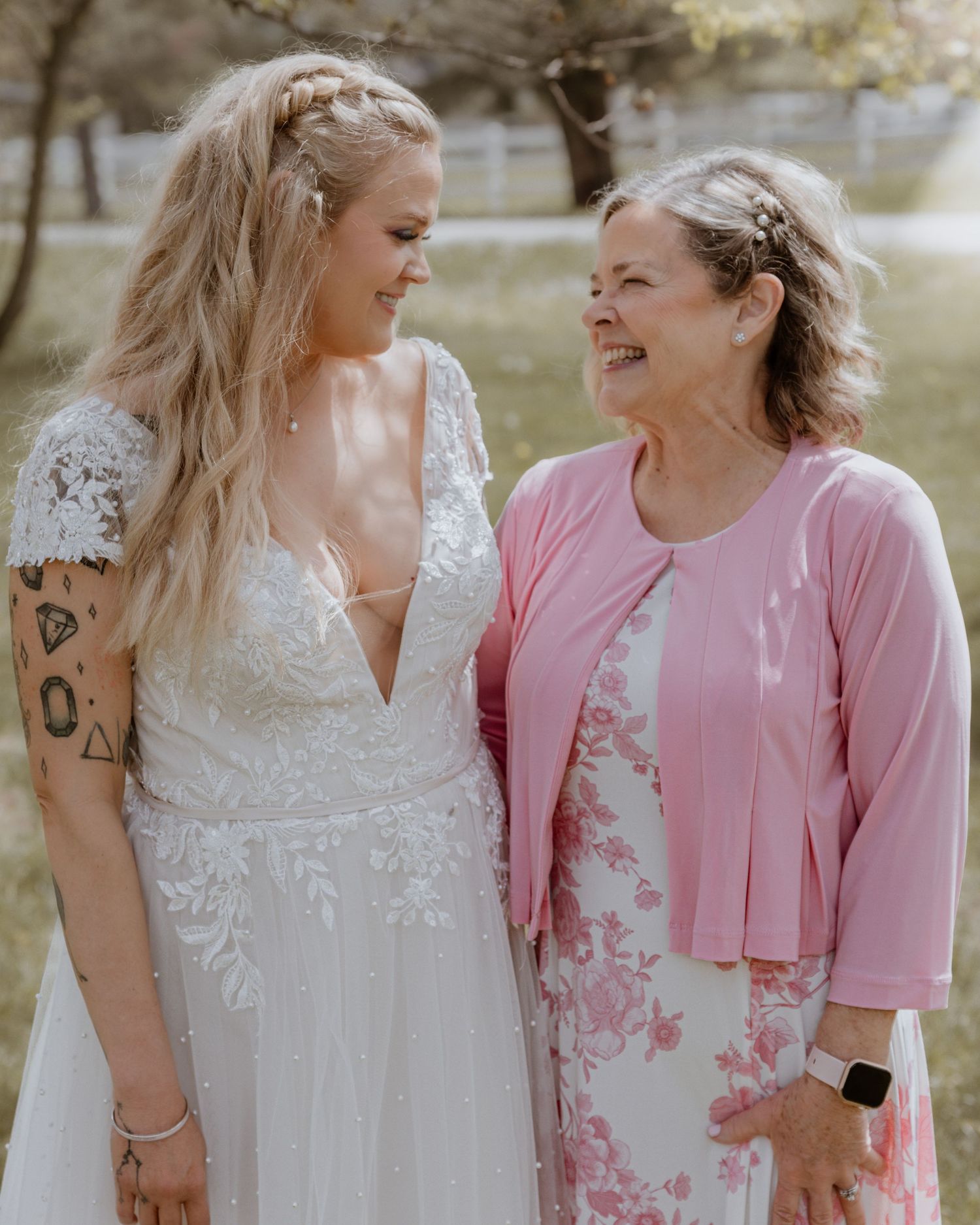 Bride in lace wedding gown smiles with older woman in pink floral dress outdoors.