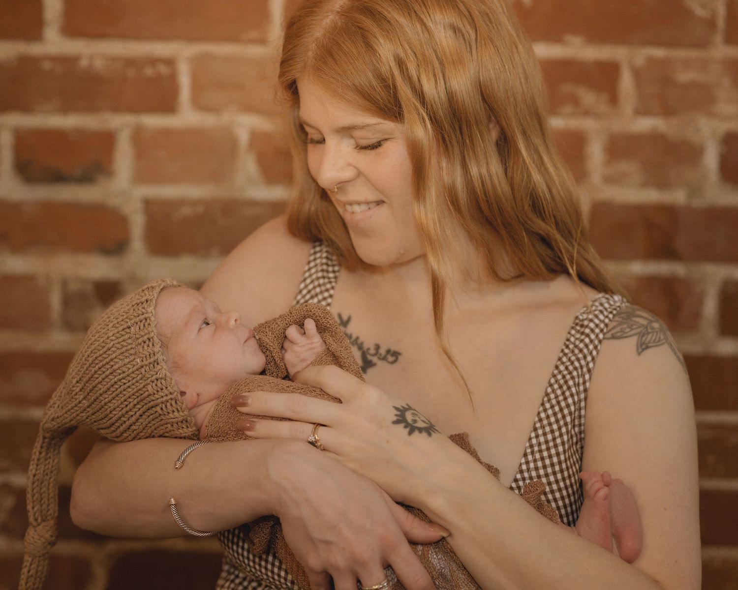 Smiling tattooed mother holding newborn baby in knit hat against brick wall.